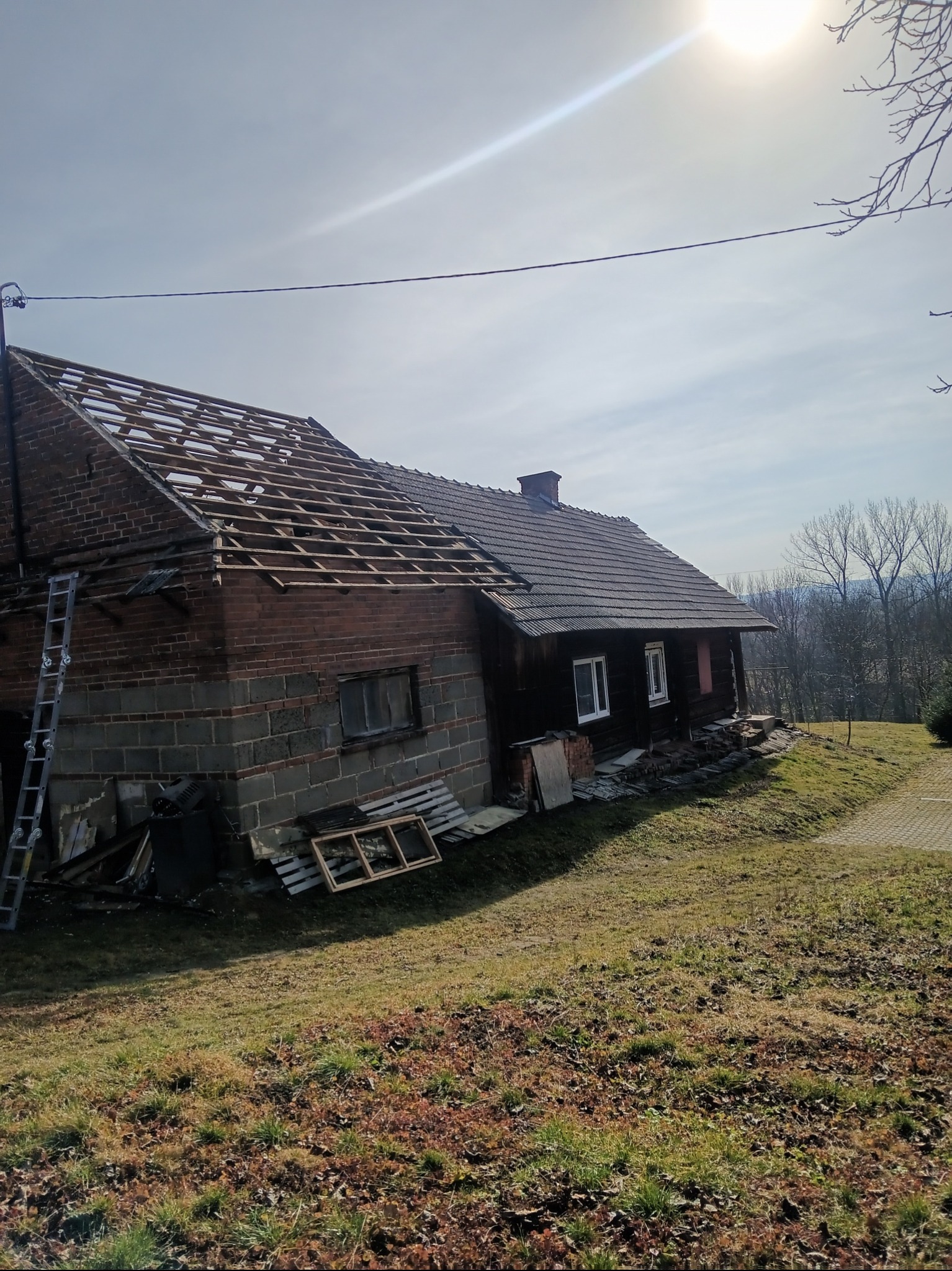 Dom z częściowo zdjętym dachem, widoczna konstrukcja więźby dachowej, drabina oparta o ścianę, teren trawiasty.