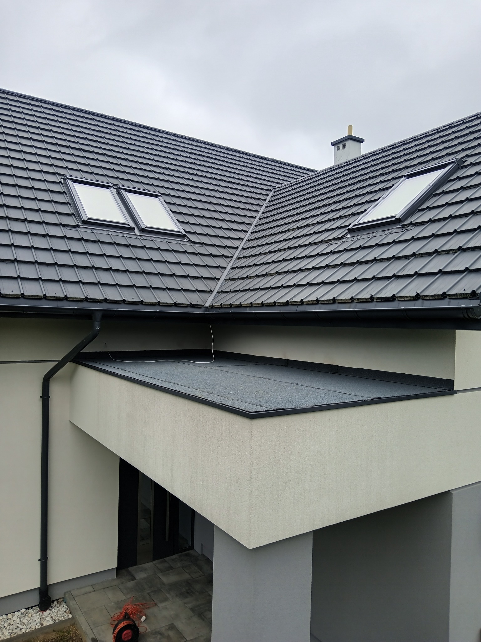 Czarny dach z dachówką i oknami dachowymi, widok z góry. Rynna pionowa po lewej stronie. Fragment elewacji budynku w neutralnym kolorze.