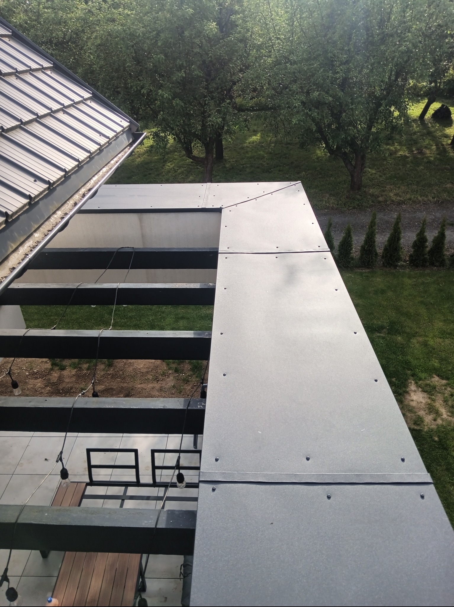 Nowoczesna konstrukcja dachu z szarej blachy na tle zieleni. Widoczne elementy mocujące i fragment rynny. Ujęcie z góry.