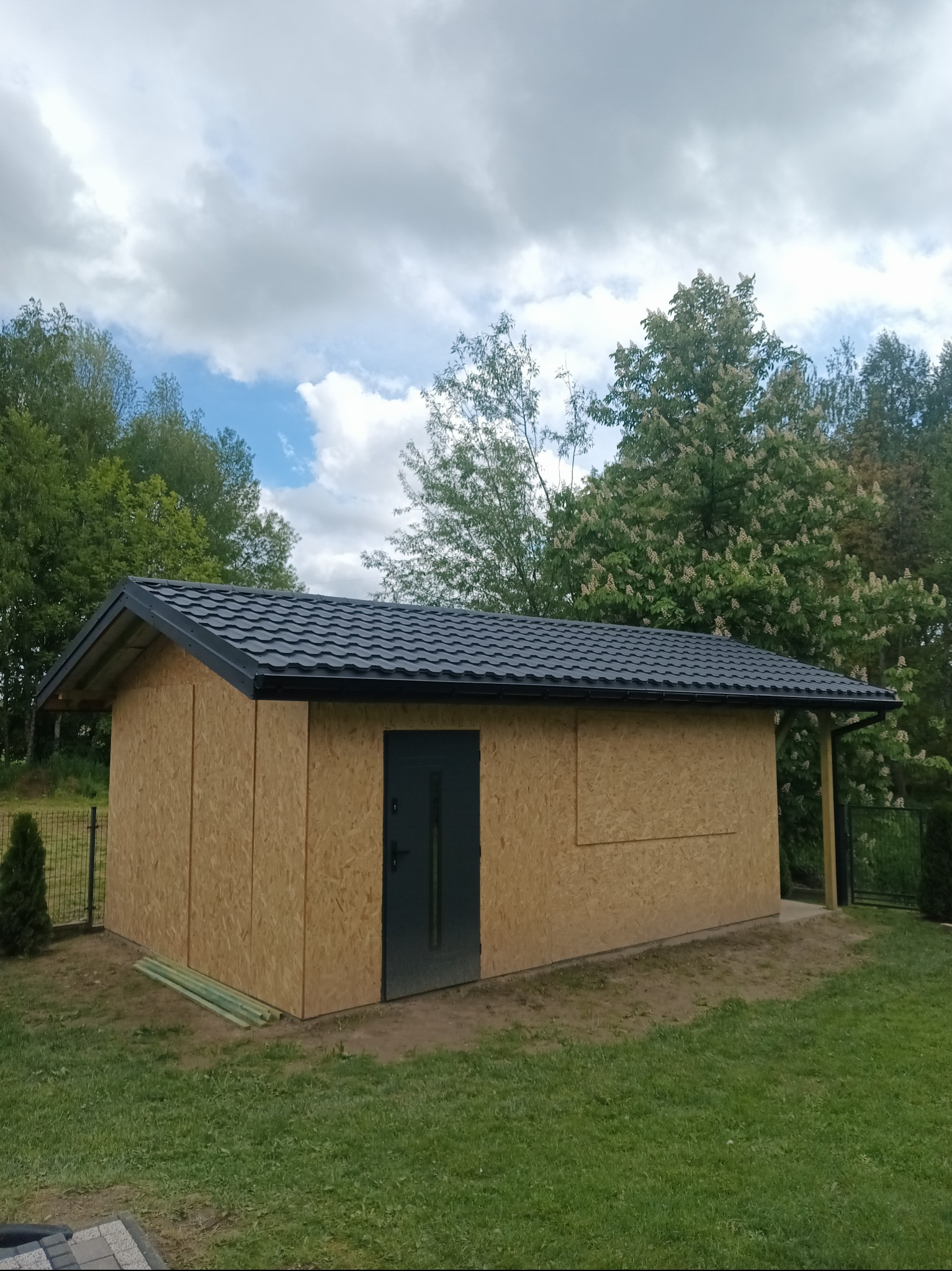 Nowy, mały budynek gospodarczy z dachem z blachy trapezowej, obity płytami OSB. Na ścianie widoczne niezabezpieczone krawędzie płyt. Drzwi wejściowe stalowe, grafitowe.