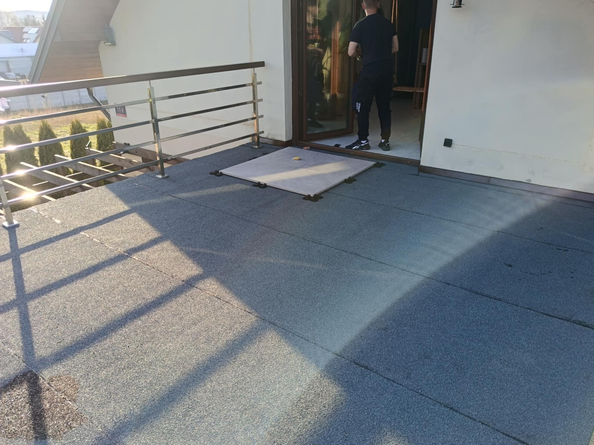 Montaż płytek na balkonie z widocznymi elementami mocującymi. Osoba w tle wchodzi do pomieszczenia. Metalowa balustrada balkonu w słoneczny dzień.