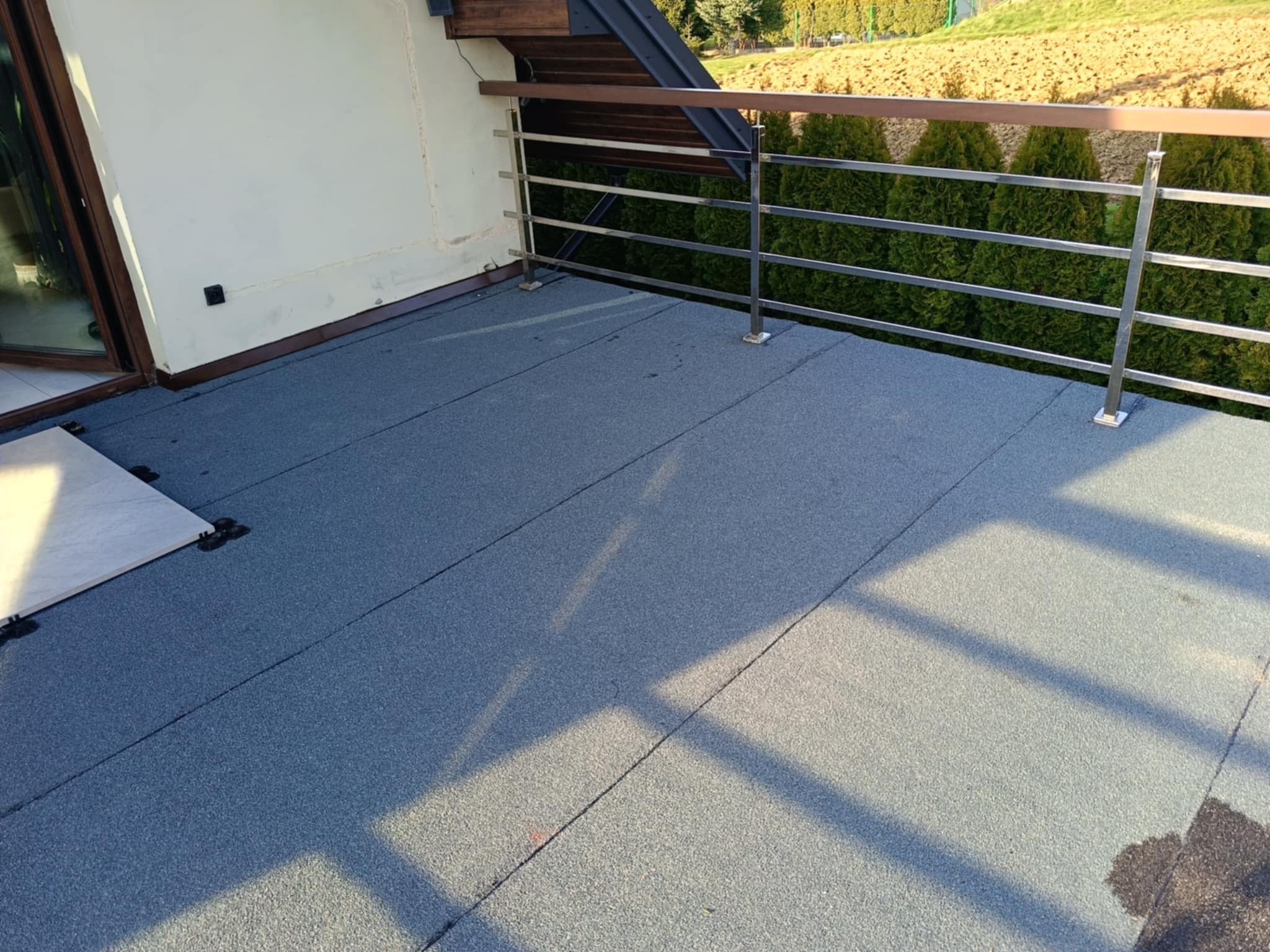 Szary dach balkonu z metalową balustradą i widokiem na zieleń. Nowoczesne wykończenie powierzchni dachu, widoczne łączenia materiału.