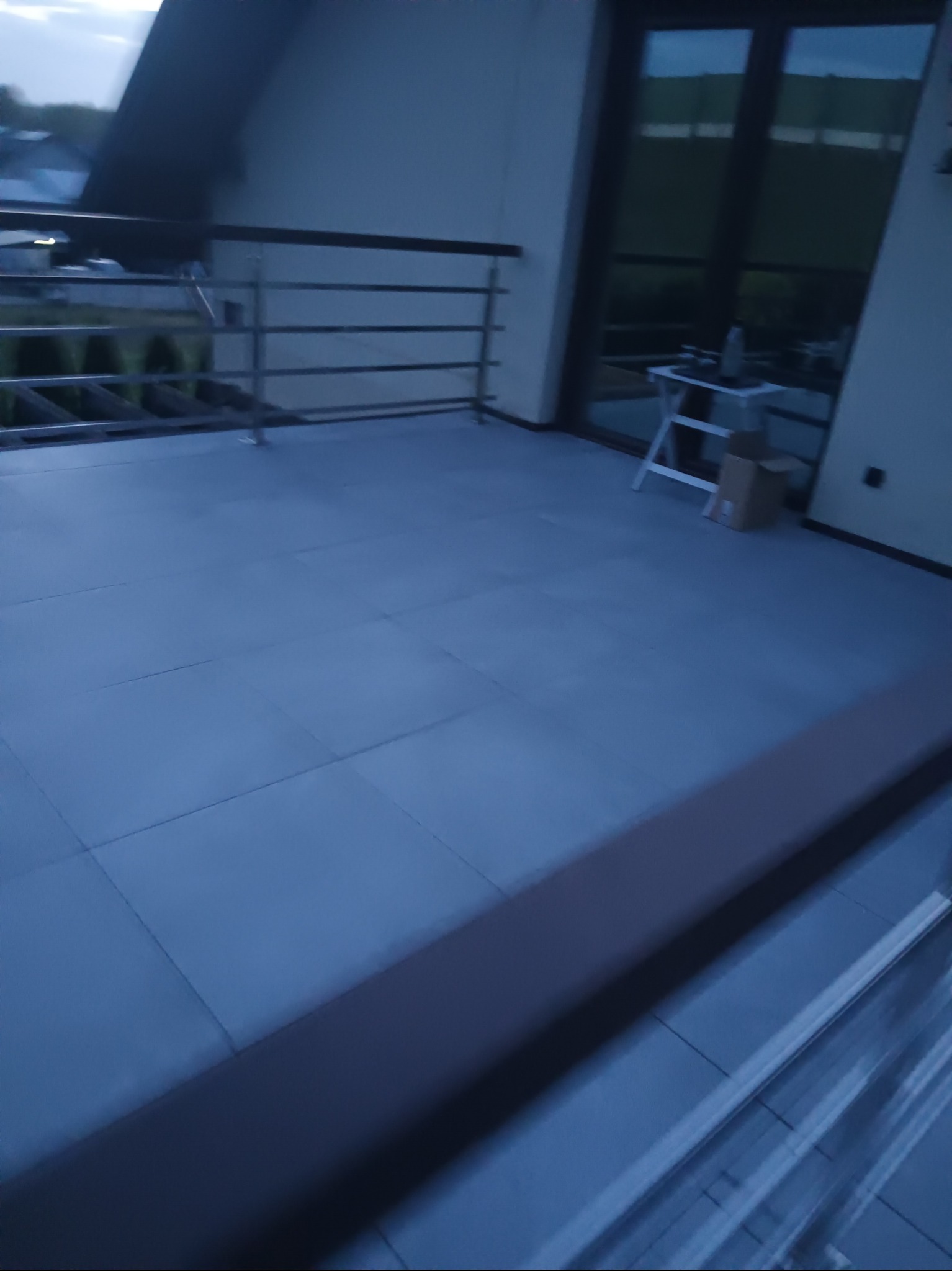 Nowoczesny balkon z szarą podłogą z płytek, metalową balustradą i dużym oknem balkonowym. Widoczne są elementy wyposażenia wnętrza.