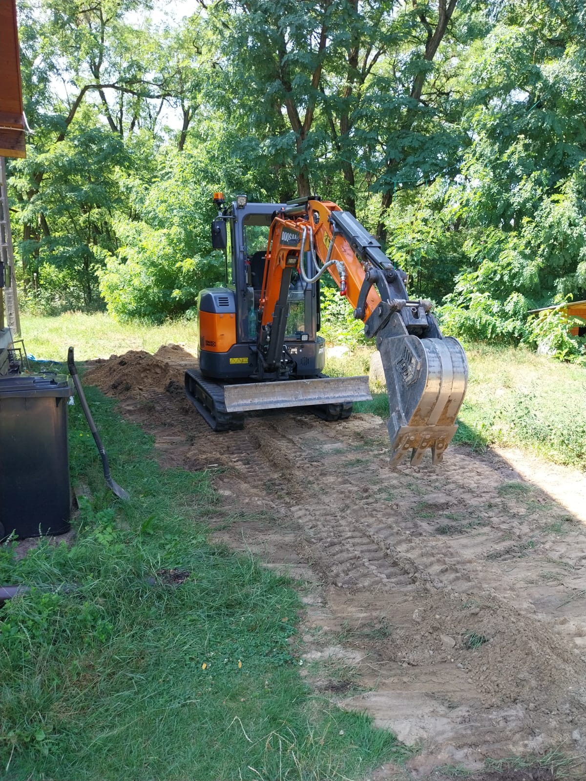 Pomarańczowa mini koparka na gąsienicach, z łyżką skierowaną w dół, przygotowuje teren pod budowę, widoczne ślady gąsienic w ziemi i drzewa w tle.