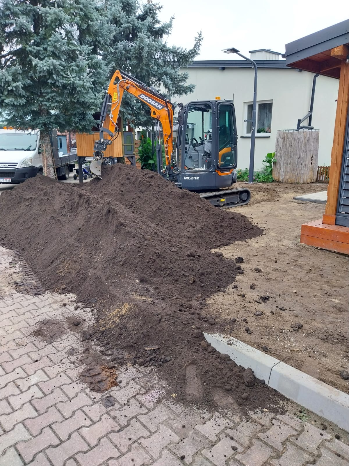 Pomarańczowa mini koparka Doosan DK 27Z na placu budowy, obok sterta świeżo wykopanej ciemnej ziemi, w tle budynek mieszkalny i drzewa.