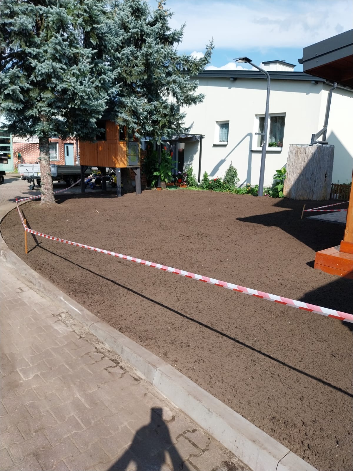 Przygotowany teren pod trawnik lub ogród, wyrównana ziemia ogrodzona taśmą ostrzegawczą, w tle budynek mieszkalny i domek na drzewie.