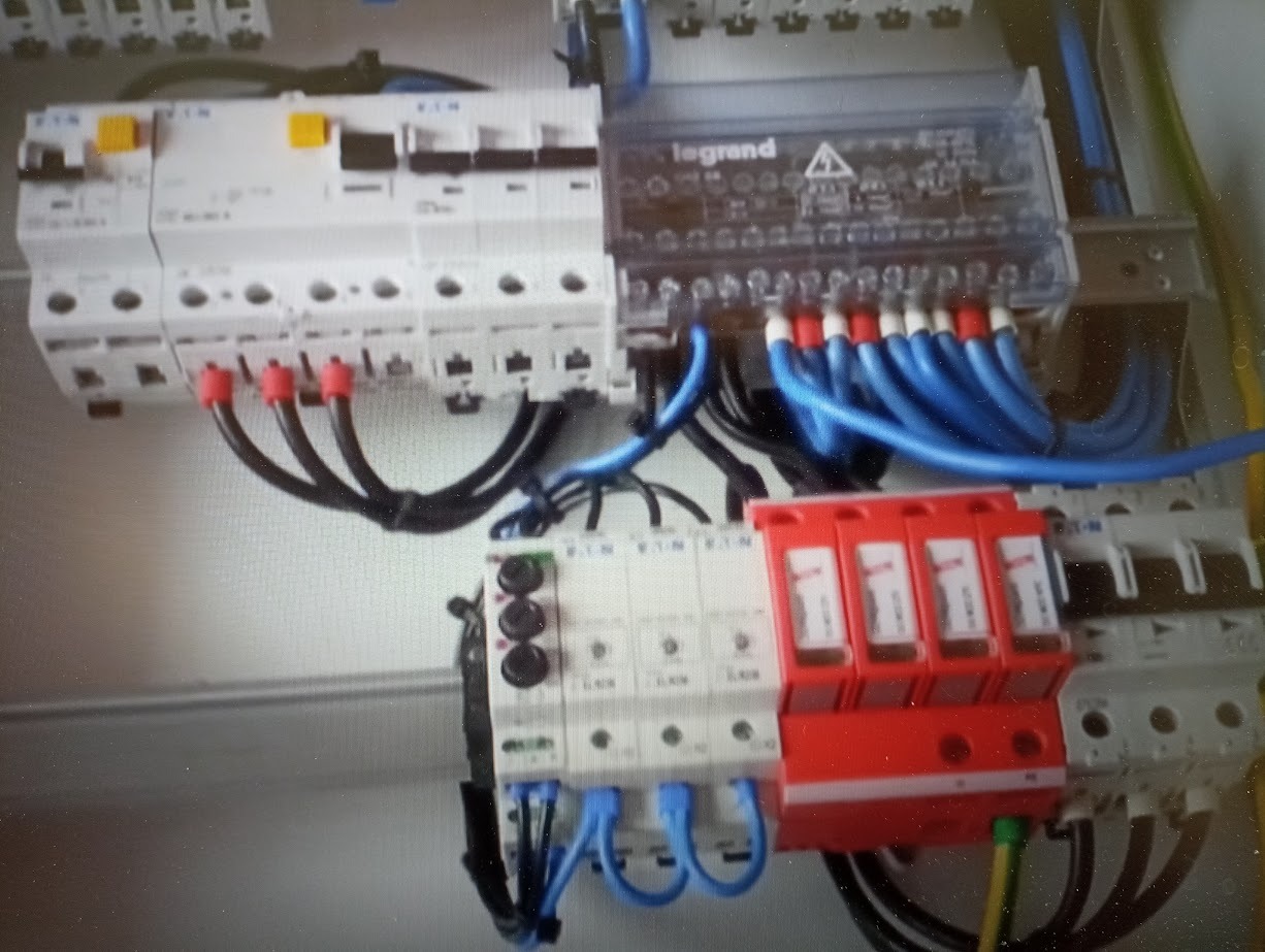 Tablica rozdzielcza z zainstalowanymi zabezpieczeniami przepięciowymi, wyłącznikami i przewodami w kolorach niebieskim i czarnym, widoczne elementy marki Legrand i Eaton.