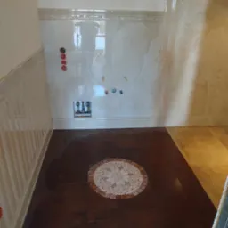 Łazienka w trakcie prac wykończeniowych: brązowe płytki podłogowe z dekoracyjną mozaiką w kształcie rozety, marmurowe płytki ścienne z ozdobną listwą i panelami, przygotowane punkty instalacyjne.