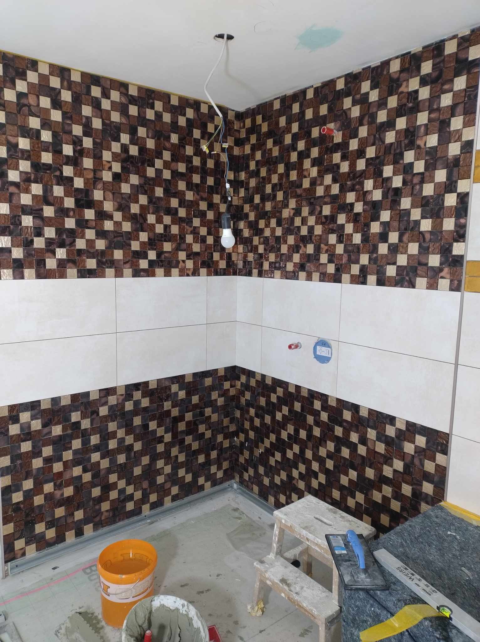 Ściana łazienki w trakcie wykańczania: mozaika z brązowo-beżowych kwadratów, przedzielona pasem białych płytek, widoczne narzędzia glazurnicze i wiadro z zaprawą.