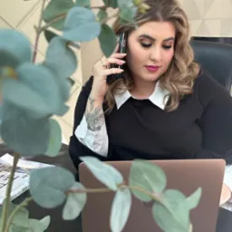 Kobieta z tatuażem na przedramieniu rozmawia przez telefon w biurze, na biurku laptop i katalogi.