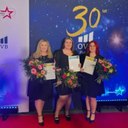 Trzy kobiety w czarnych sukienkach stoją na czerwonym dywanie, trzymając bukiety kwiatów i dyplomy na tle baneru OVB z okazji 30-lecia firmy.