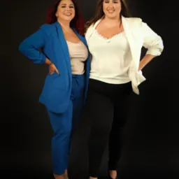 Dwie uśmiechnięte kobiety w eleganckich strojach biznesowych, jedna w niebieskim garniturze i druga w białej marynarce, stoją obok siebie na czarnym tle.