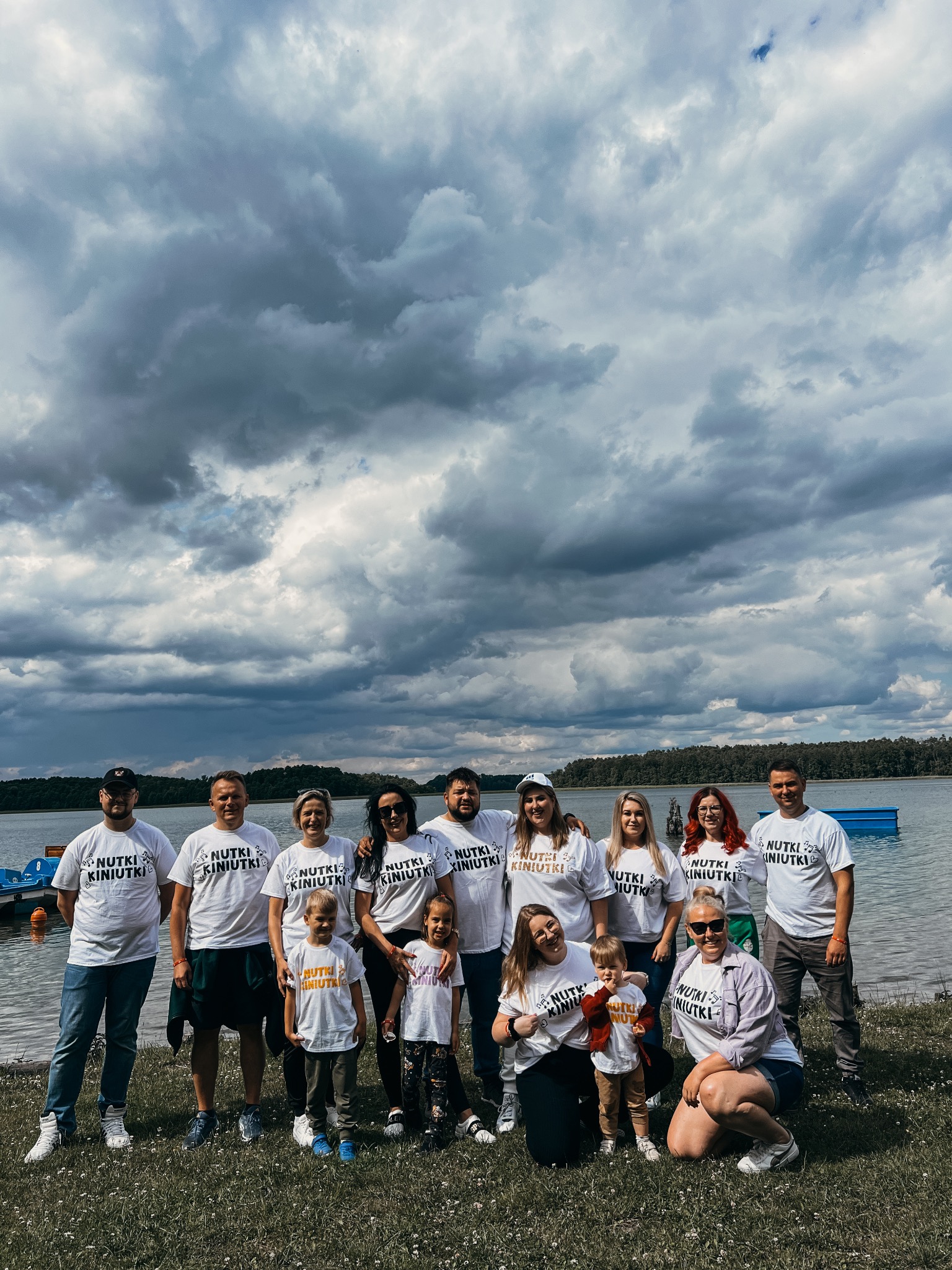 Grupa kilkunastu osób w białych koszulkach z napisem 'Nutki Kiniutki' pozuje na tle jeziora i pochmurnego nieba.