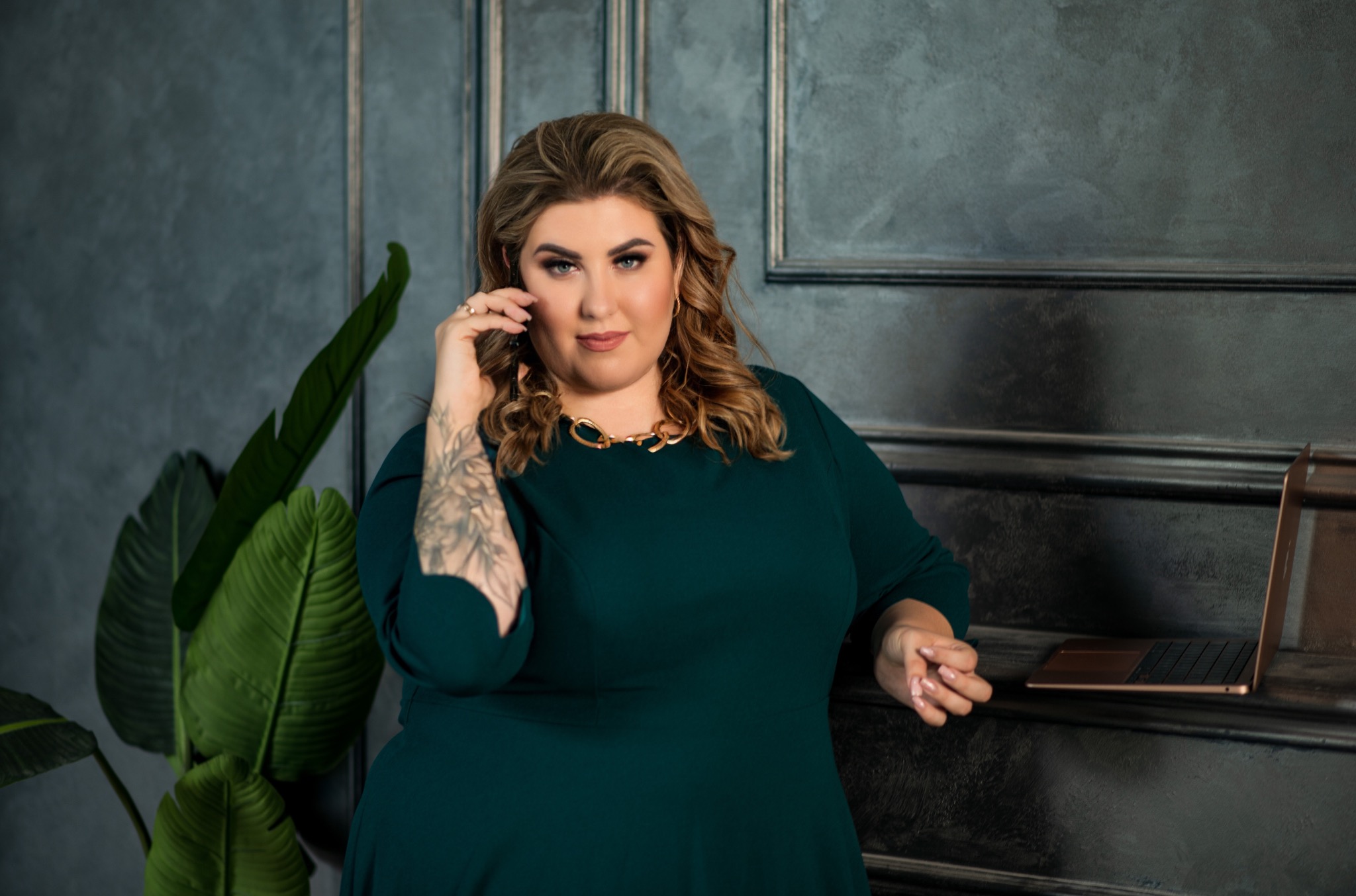 Kobieta w zielonej sukience rozmawia przez telefon, opierając się o ciemną ścianę z laptopem i rośliną w tle.