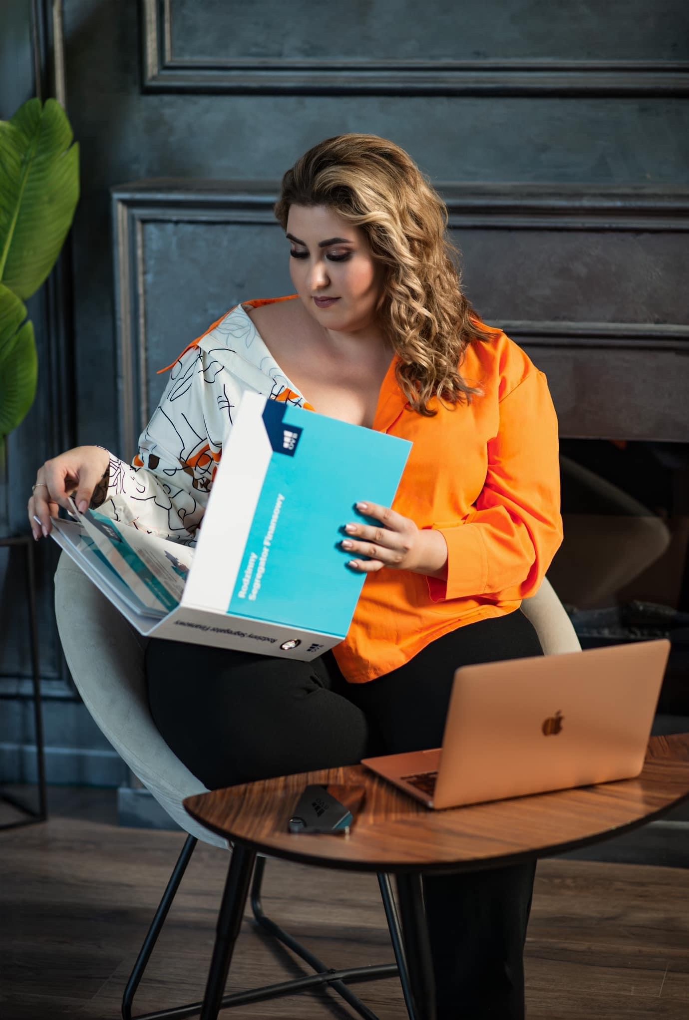 Kobieta w pomarańczowej bluzce przegląda segregator finansowy siedząc na krześle obok laptopa na małym stoliku.