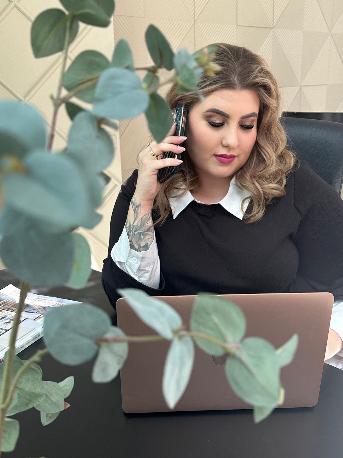 Kobieta z tatuażem na przedramieniu rozmawia przez telefon w biurze, na biurku laptop i katalogi.