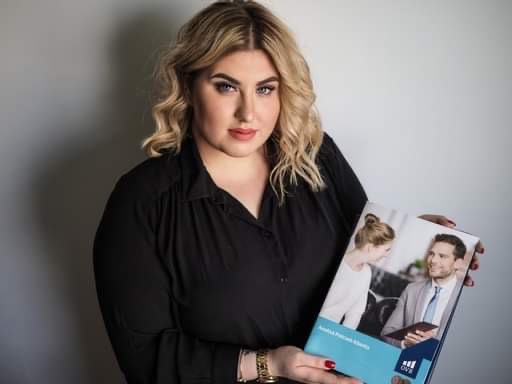 Kobieta z blond lokami prezentuje otwarty folder reklamowy firmy finansowej z uśmiechniętą parą na okładce i logotypem w prawym dolnym rogu.