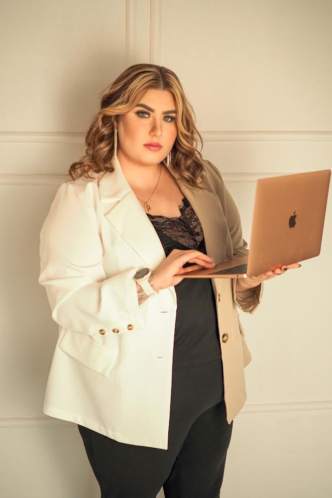 Kobieta w dwukolorowej marynarce pozuje z laptopem w dłoni, patrząc w obiektyw; widoczny zegarek na nadgarstku i długie kolczyki.