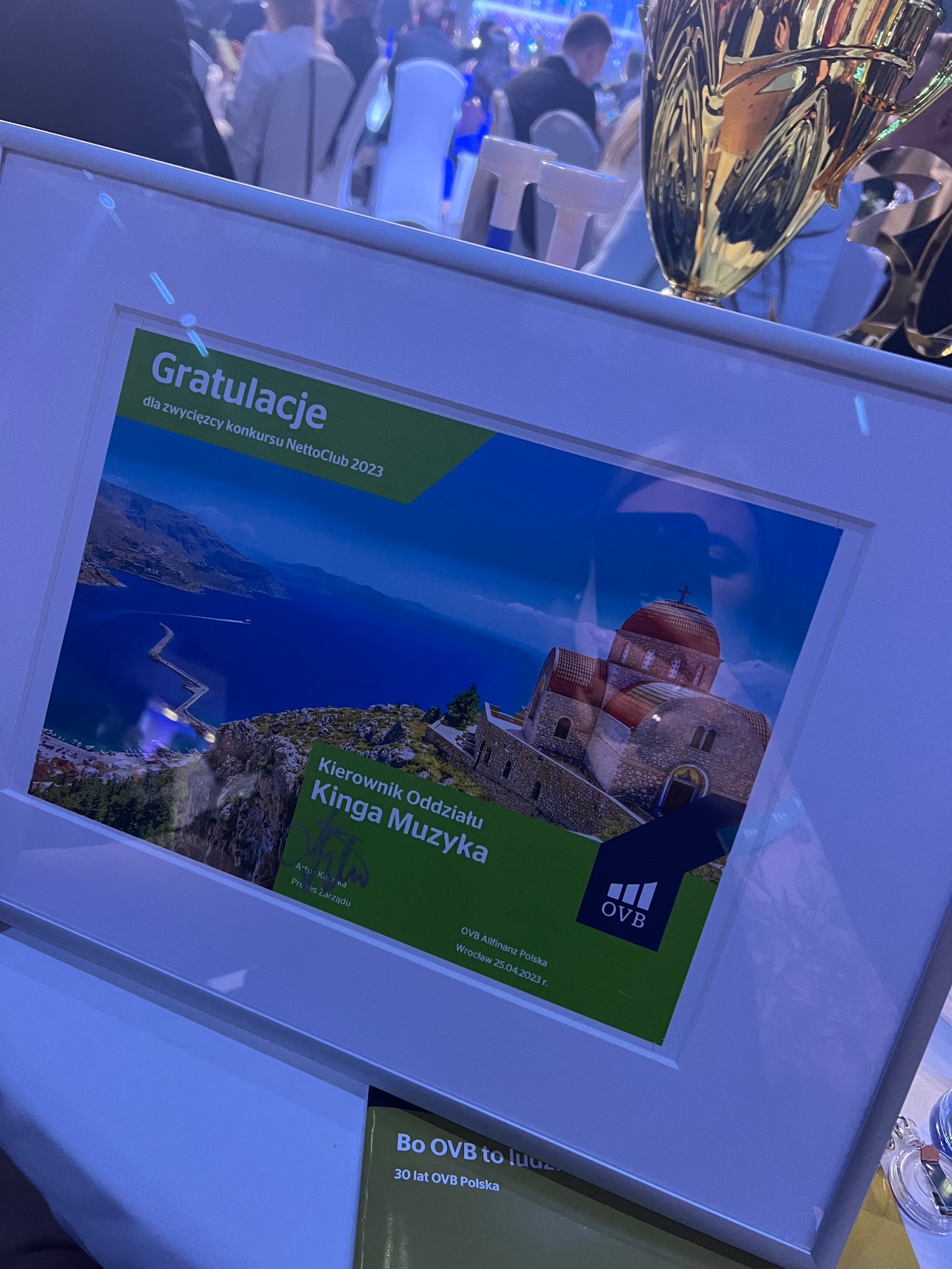 Oprawiona w ramkę grafika z gratulacjami dla Kingi Muzyki, Kierownika Oddziału, za wygraną w konkursie NettoClub 2023, z widokiem na grecką wyspę z kościołem i długim molo, obok złoty puchar.