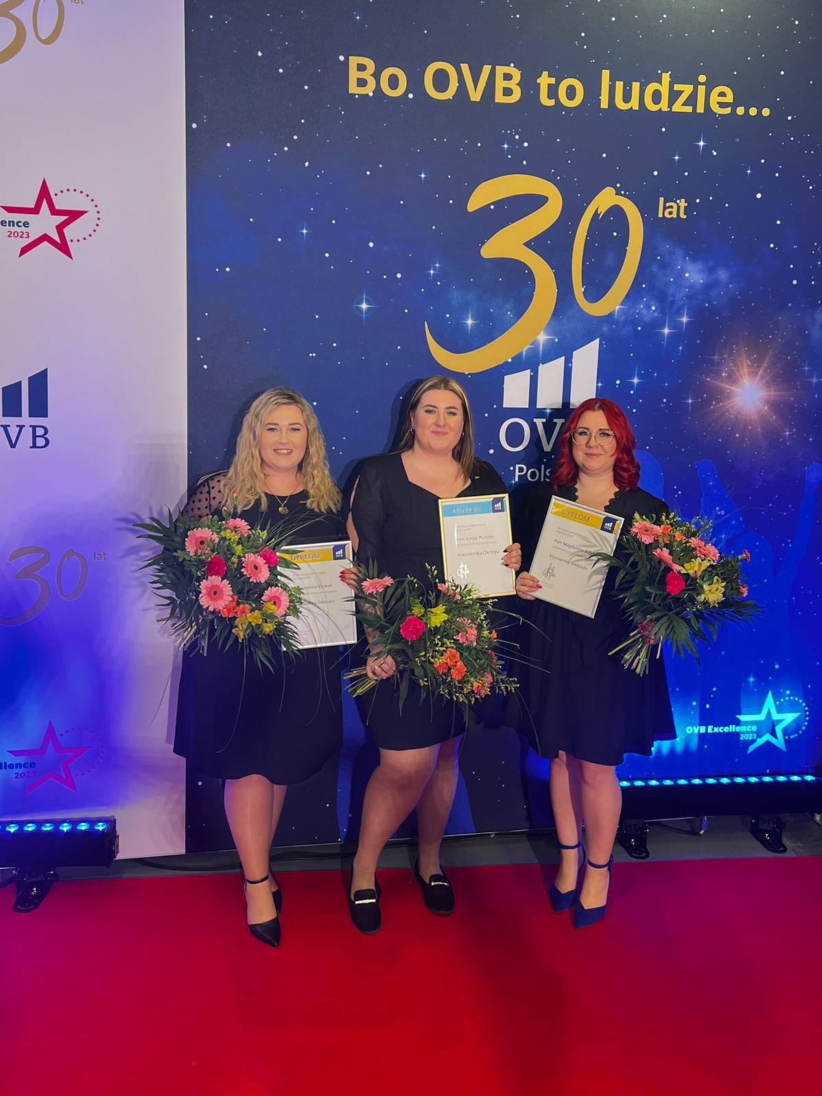 Trzy kobiety w czarnych sukienkach stoją na czerwonym dywanie, trzymając bukiety kwiatów i dyplomy na tle baneru OVB z okazji 30-lecia firmy.