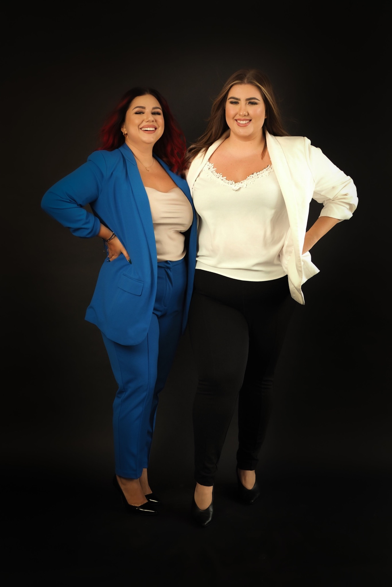 Dwie uśmiechnięte kobiety w eleganckich strojach biznesowych, jedna w niebieskim garniturze i druga w białej marynarce, stoją obok siebie na czarnym tle.