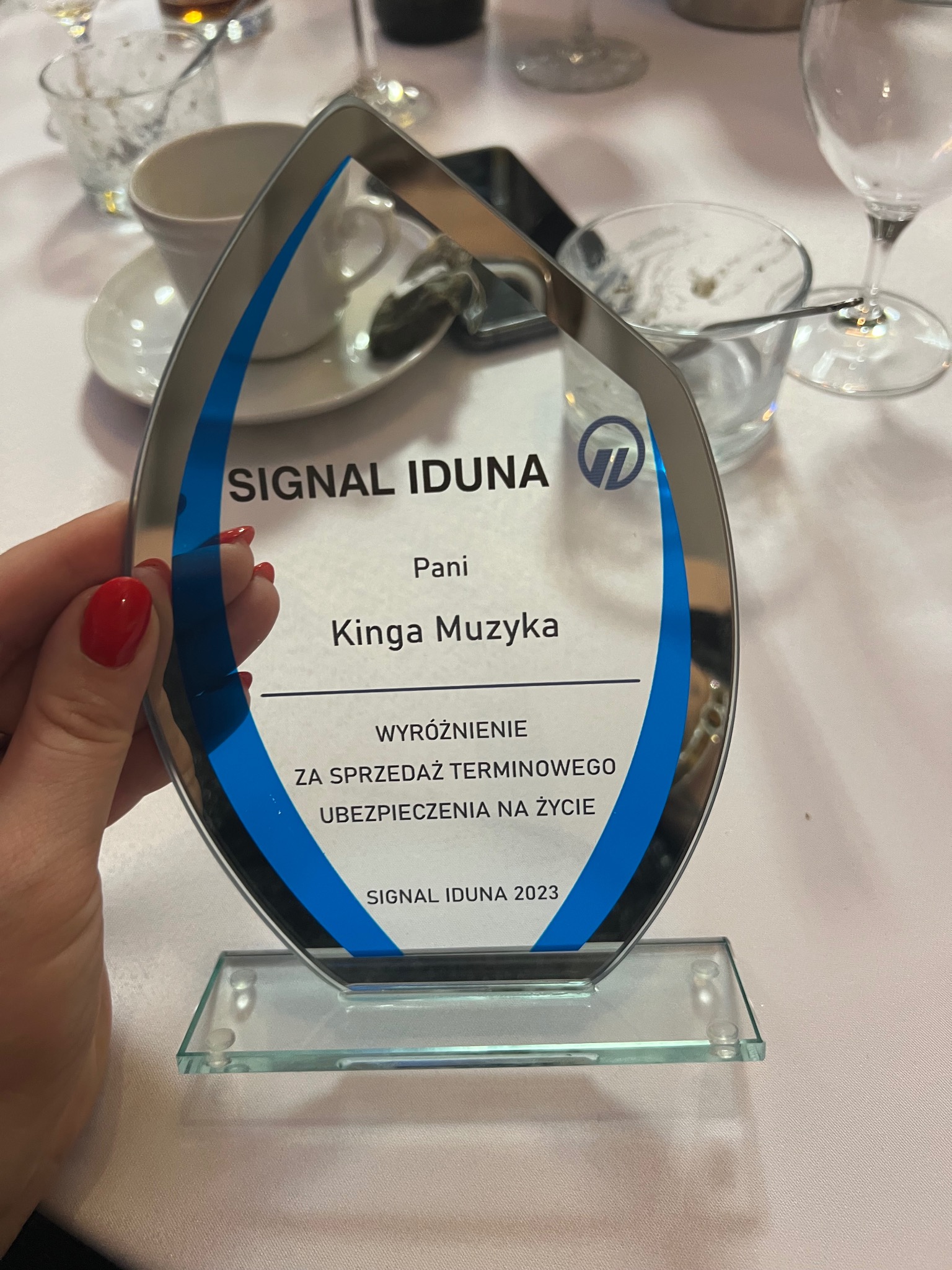 Szklana statuetka z logo Signal Iduna, wręczona Pani Kindze Muzyce za sprzedaż terminowego ubezpieczenia na życie w 2023 roku, trzymana w dłoni na tle zastawionego stołu.