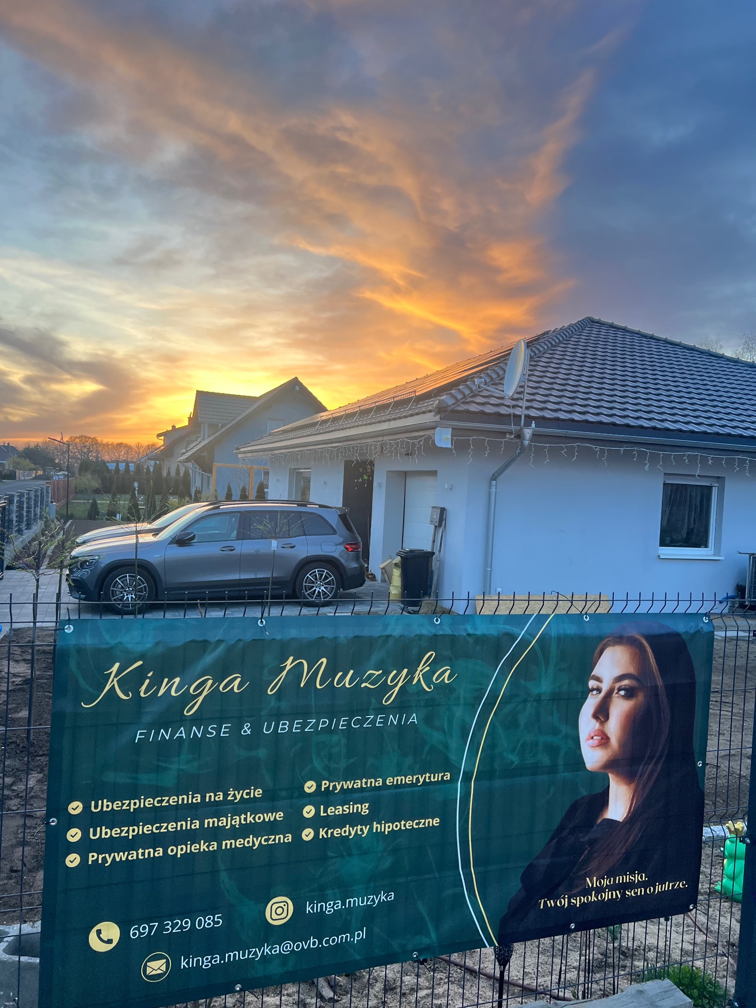 Baner reklamowy firmy finansowej i ubezpieczeniowej Kinga Muzyka na tle domu jednorodzinnego z samochodem i zachodzącym słońcem.