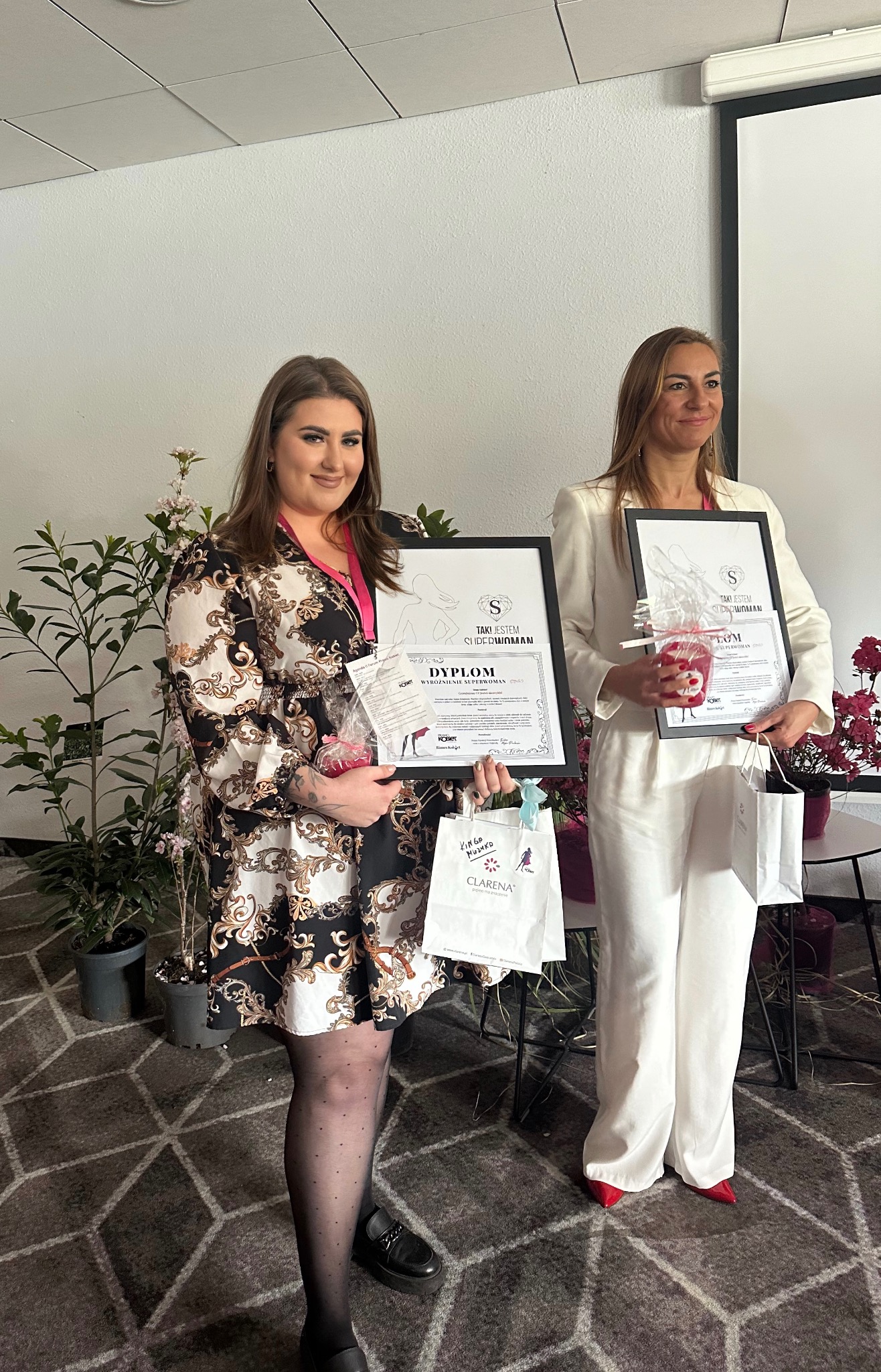Dwie kobiety prezentują dyplomy 'Superwoman' w ramkach, trzymają również upominki i torby z logo firm, na tle roślin i ściany z projektorem.