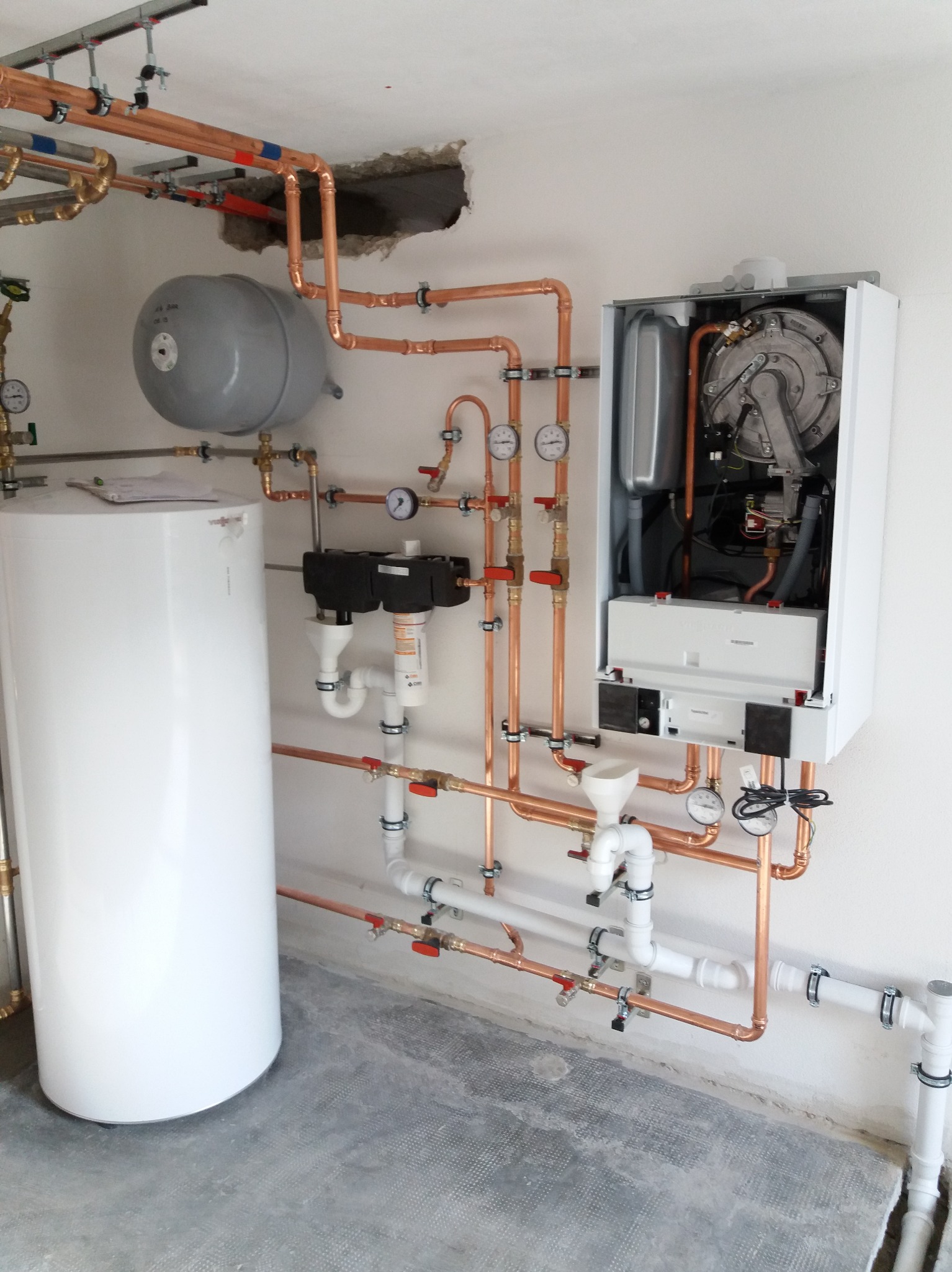 Nowoczesny, biały piec CO z otwartą obudową, widoczne wewnętrzne komponenty, połączony miedzianymi rurami z białym zasobnikiem ciepłej wody i szarym zbiornikiem wyrównawczym, zamontowany na białej...