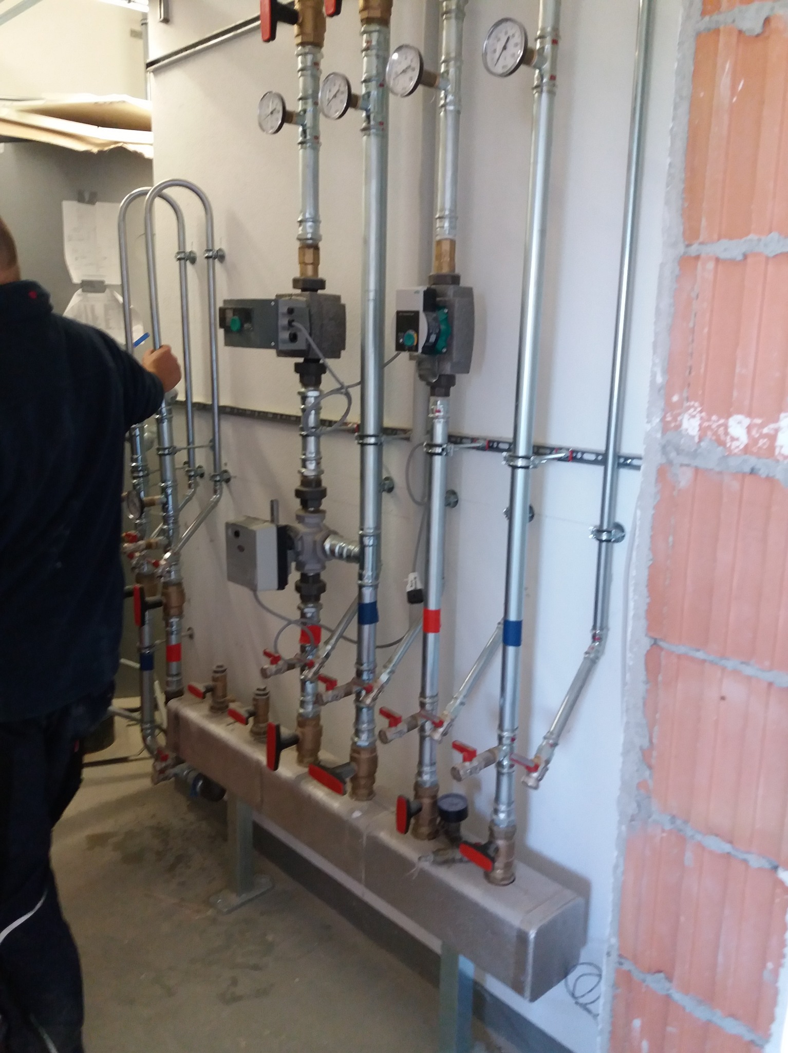 Instalacja grzewcza z rzędami rur, zaworami z czerwonymi uchwytami i manometrami, zamontowana na ścianie w surowym stanie, widoczny fragment muru z cegły.