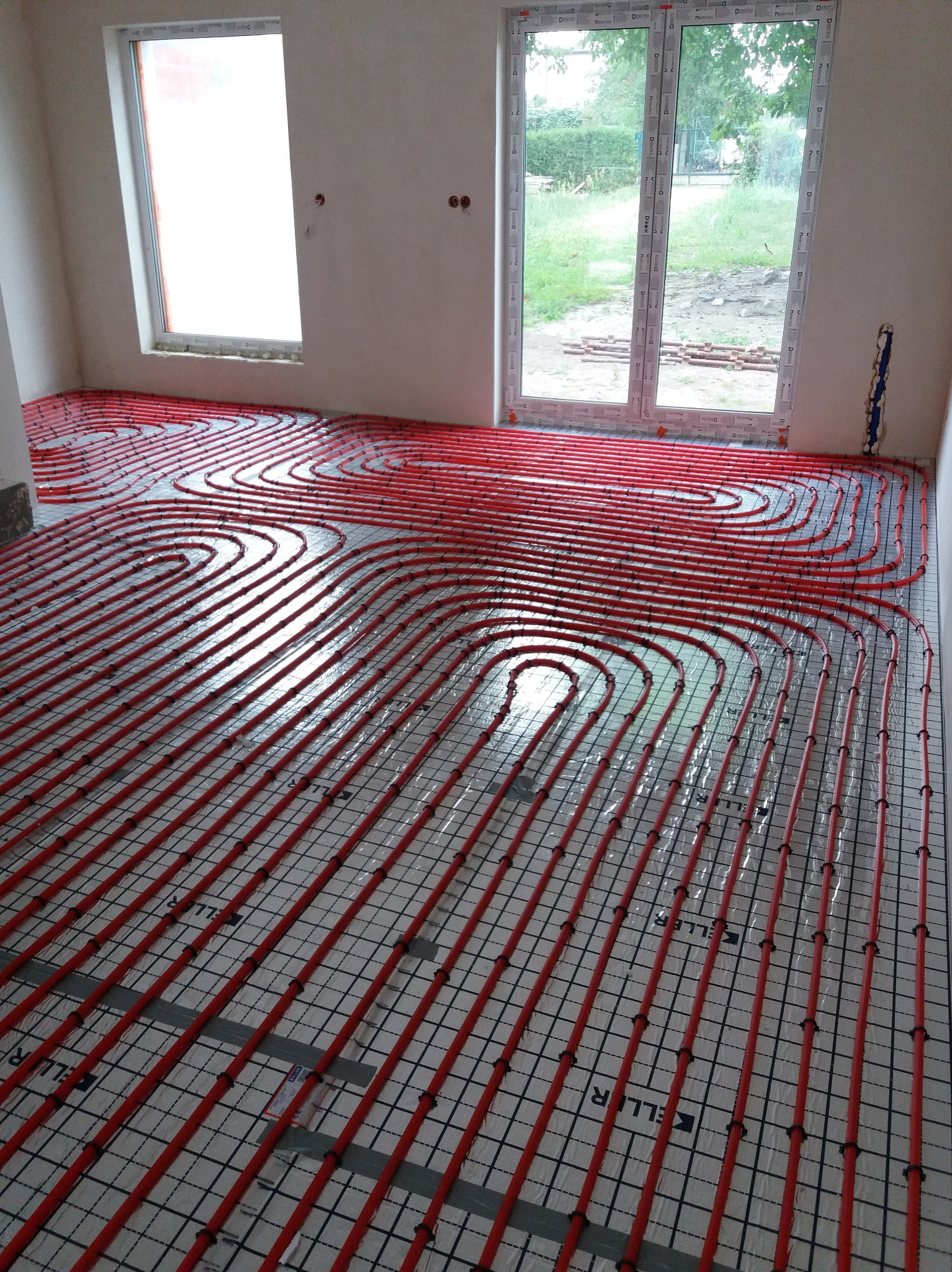 Czerwone rury ogrzewania podłogowego ułożone w pętle na czarnej siatce w jasnym pomieszczeniu z dwoma oknami.