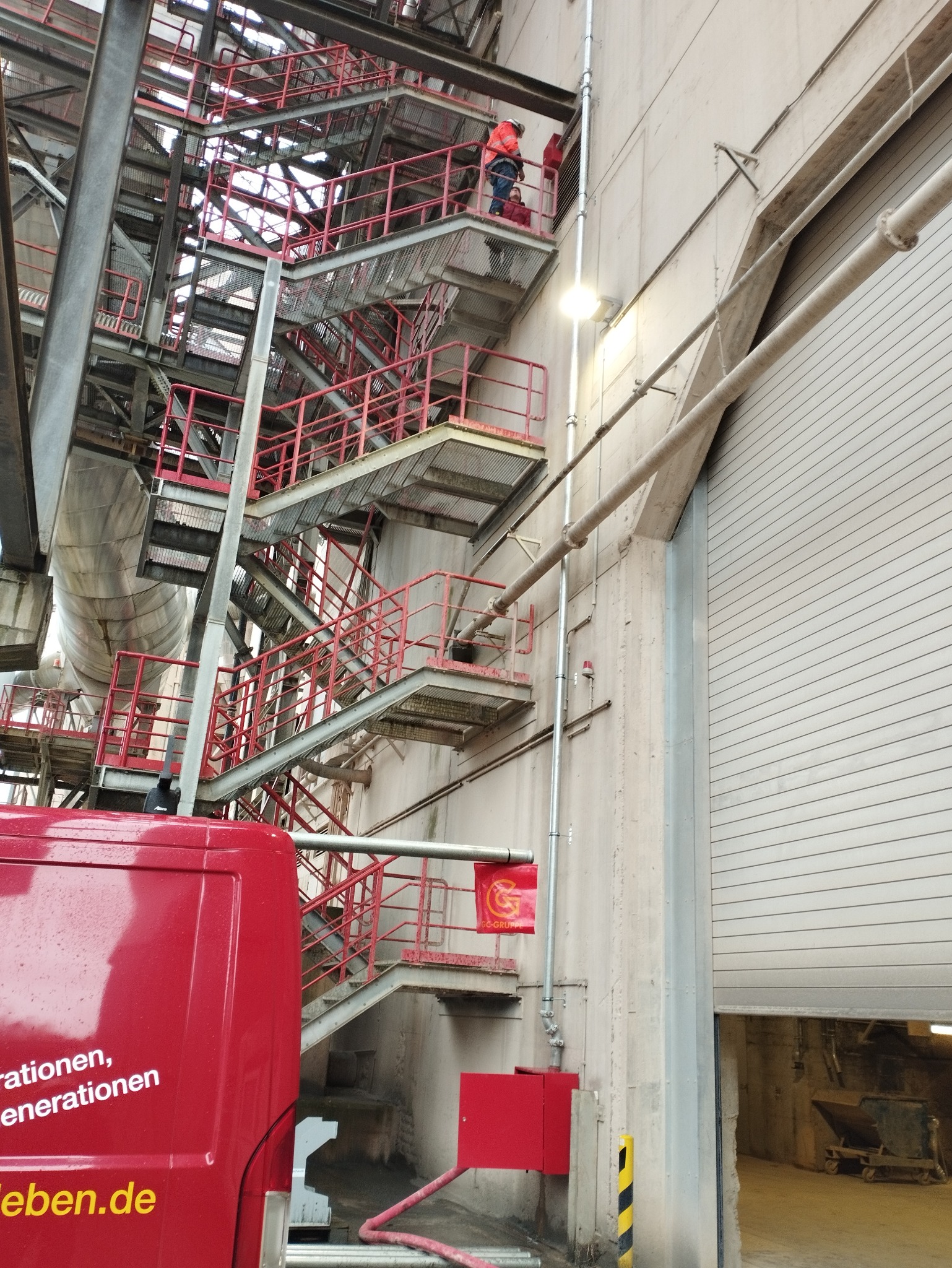 Pracownicy w pomarańczowych kombinezonach na metalowych schodach przemysłowych przy betonowej ścianie budynku, widoczna rura i brama garażowa oraz fragment czerwonego samochodu dostawczego z napisem.