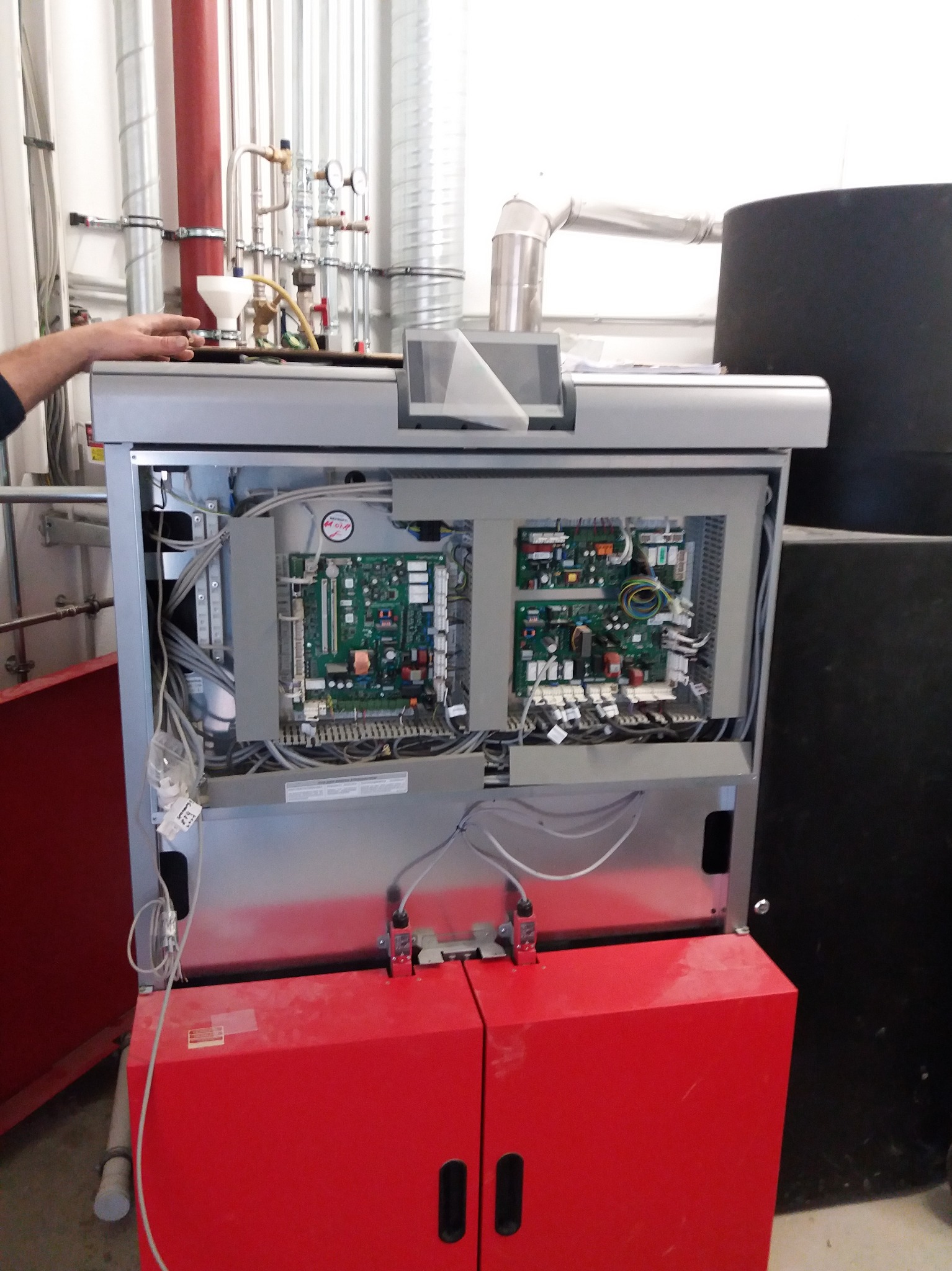 Otwarta obudowa kotła z widocznymi płytami sterującymi i okablowaniem, czerwona szafka dolna, instalacja rurowa w tle.