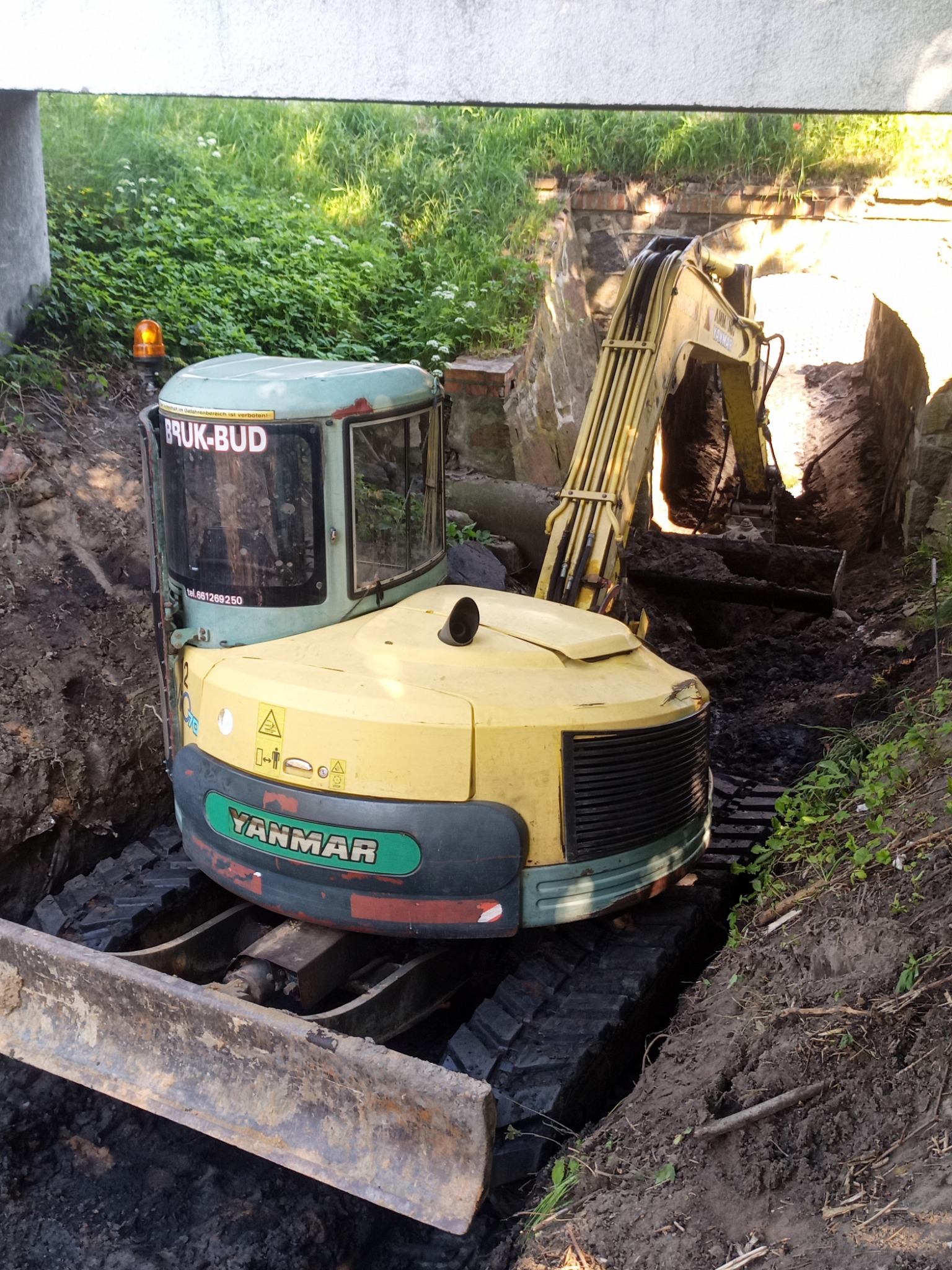Żółto-zielona minikoparka Yanmar stoi w wąskim wykopie, z widocznymi śladami gąsienic w błotnistej ziemi, na kabinie naklejka 'BRUK-BUD' i numer telefonu, tło stanowi zielona roślinność i mur...