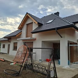 JAR-BUD JAROSŁAW CISZEK - Niedawno otynkowany dom jednorodzinny z brązowymi roletami, widoczne rusztowanie i pracownik przy elewacji, na pierwszym planie huśtawka ogrodowa.