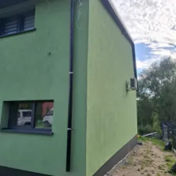 Świeżo otynkowana, zielona elewacja domu jednorodzinnego z czarną rynną i widocznym klimatyzatorem na bocznej ścianie, z elementami wykończenia przy gruncie.