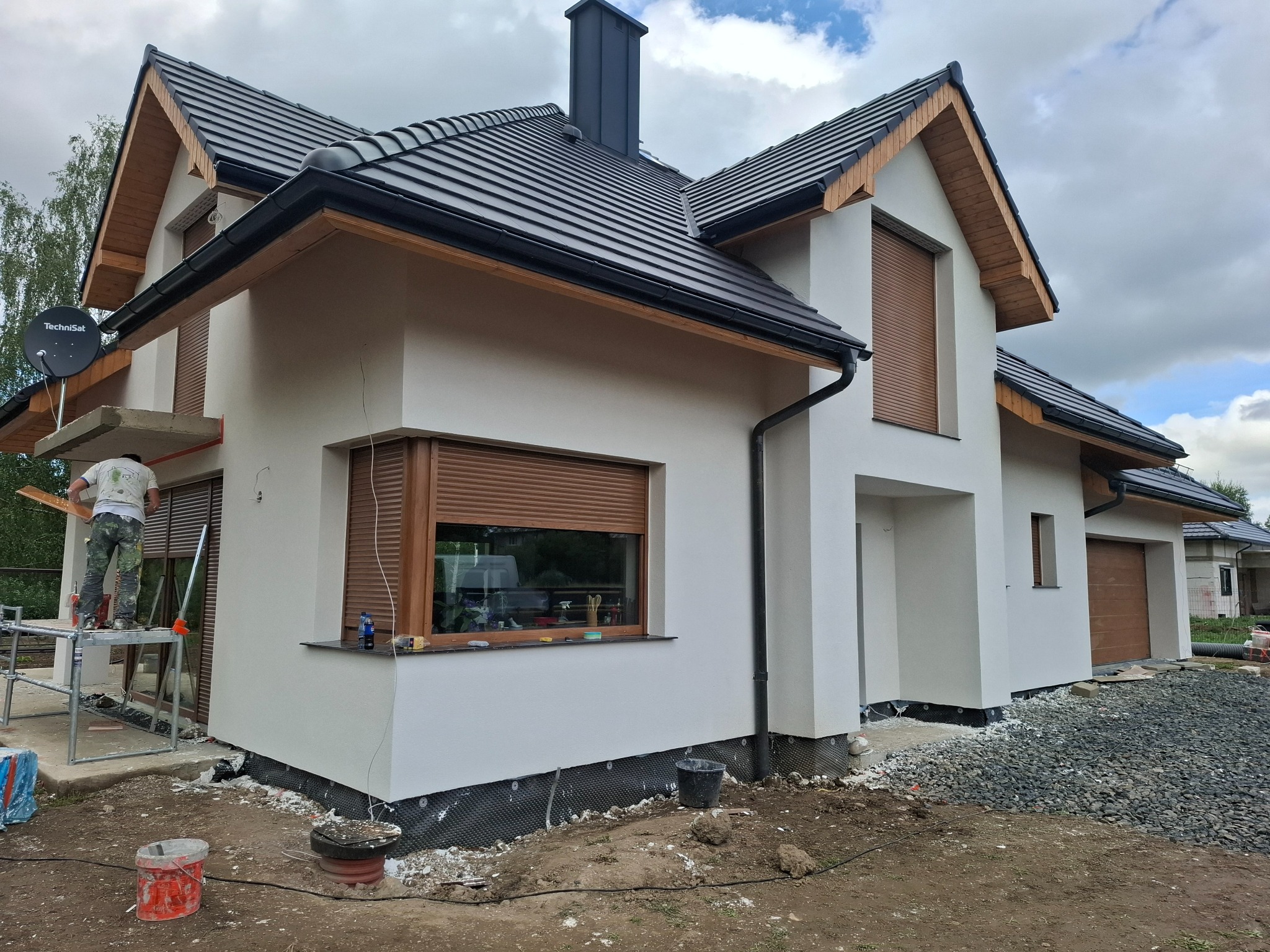 Boczny widok domu jednorodzinnego w trakcie prac wykończeniowych elewacji, widoczny pracownik na rusztowaniu i przygotowane podłoże wokół budynku.