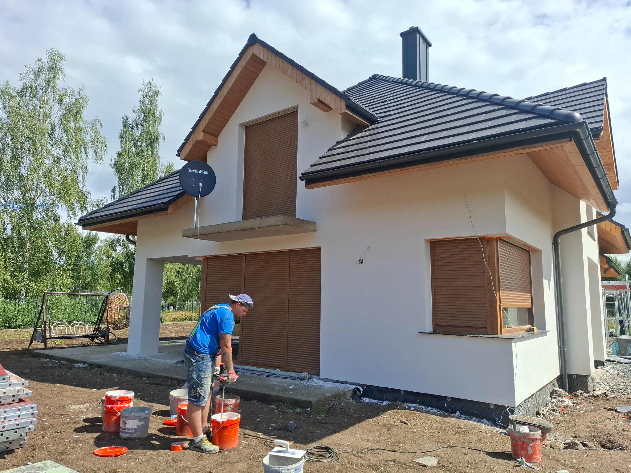 Pracownik budowlany miesza zaprawę w wiadrze przed budynkiem z jasną elewacją, roletami zewnętrznymi i anteną satelitarną na dachu, widoczny bałagan na placu budowy.