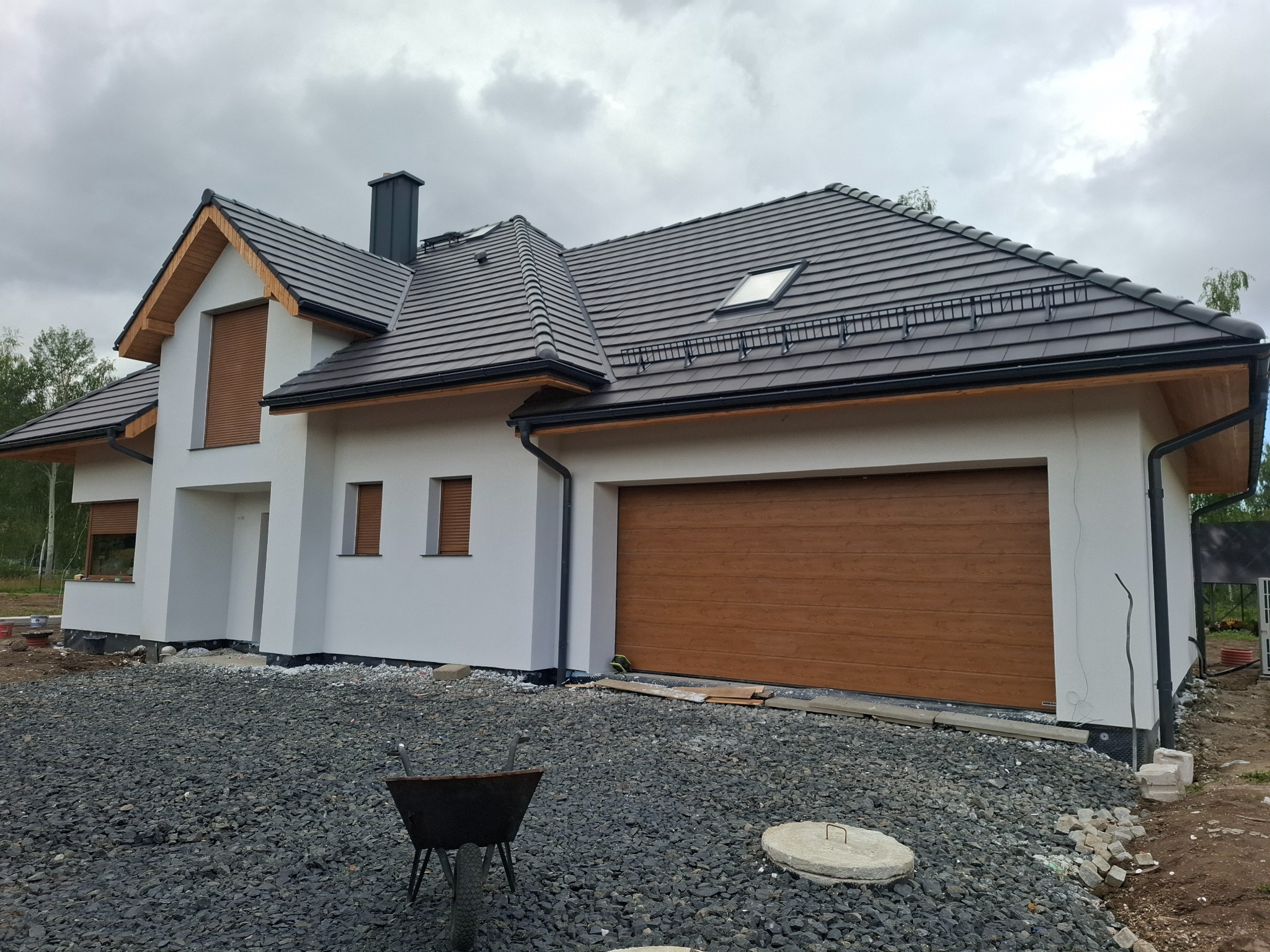 Nowoczesny dom jednorodzinny z brązową bramą garażową i grafitową dachówką, widok z zewnątrz na etapie wykończenia terenu, z widocznym wózkiem budowlanym na żwirowym podłożu.