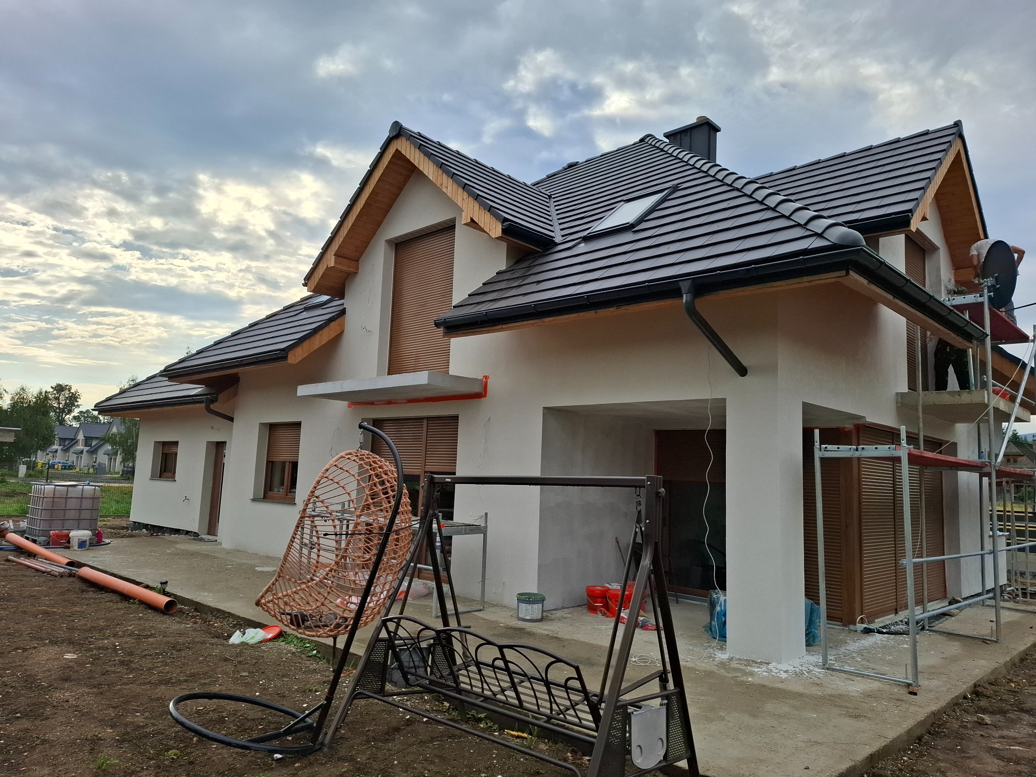 Niedawno otynkowany dom jednorodzinny z brązowymi roletami, widoczne rusztowanie i pracownik przy elewacji, na pierwszym planie huśtawka ogrodowa.