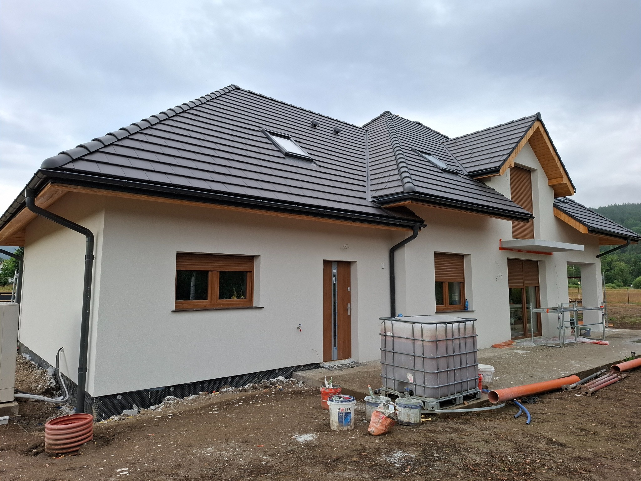 Nowy dom z jasną elewacją i brązową dachówką, widoczne okna dachowe, rynny, oraz materiały budowlane na posesji w trakcie prac wykończeniowych.