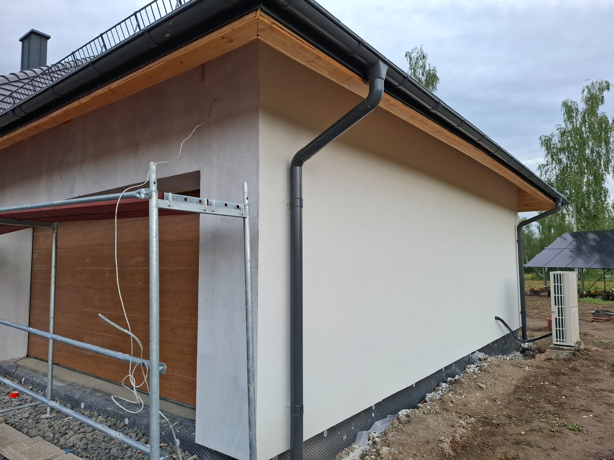 Fragment budynku z ocieploną elewacją w trakcie wykończenia, widoczne rusztowanie, orynnowanie i pompa ciepła na zewnątrz.