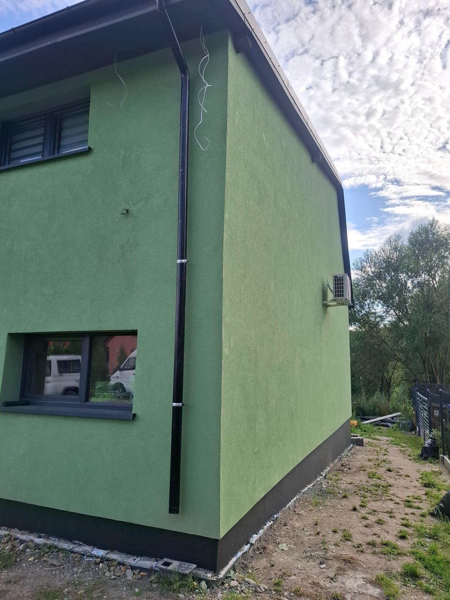 Świeżo otynkowana, zielona elewacja domu jednorodzinnego z czarną rynną i widocznym klimatyzatorem na bocznej ścianie, z elementami wykończenia przy gruncie.