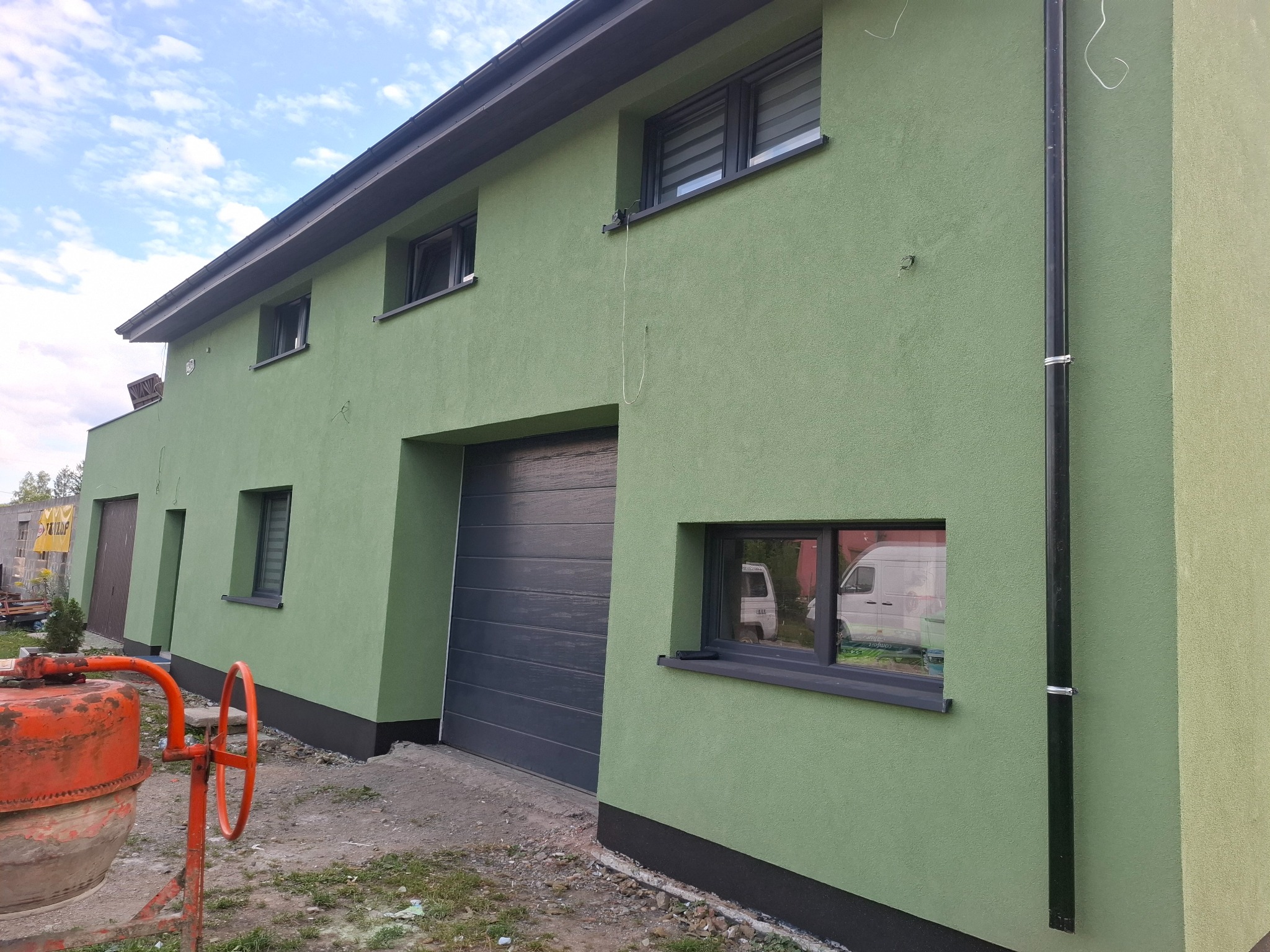Budynek z zieloną elewacją o nierównej fakturze, widoczne szare okna, brama garażowa i pomarańczowa betoniarka z przodu, w oknie odbija się biały bus.