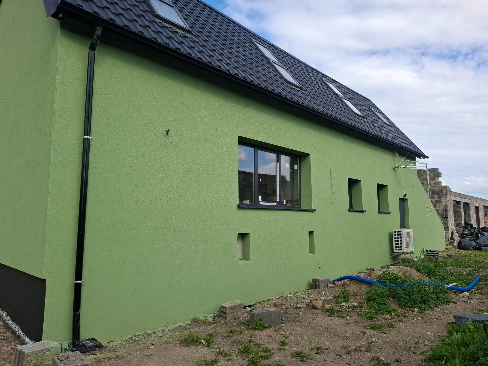 Świeżo otynkowany, pistacjowy dom jednorodzinny z czarną dachówką i rynnami, widok z boku na tle błękitnego nieba, z fragmentem niedokończonej konstrukcji w tle.