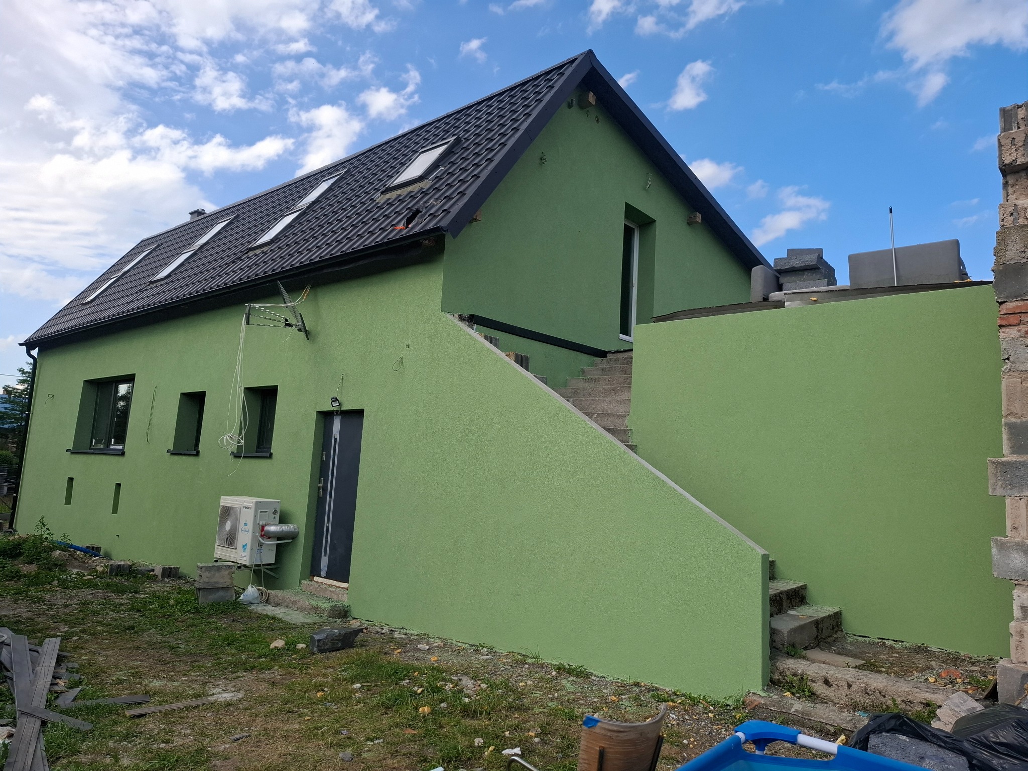 Dom z nową, jednolitą, zieloną elewacją, widoczne schody zewnętrzne, okna dachowe i klimatyzator, teren wokół domu częściowo uporządkowany.