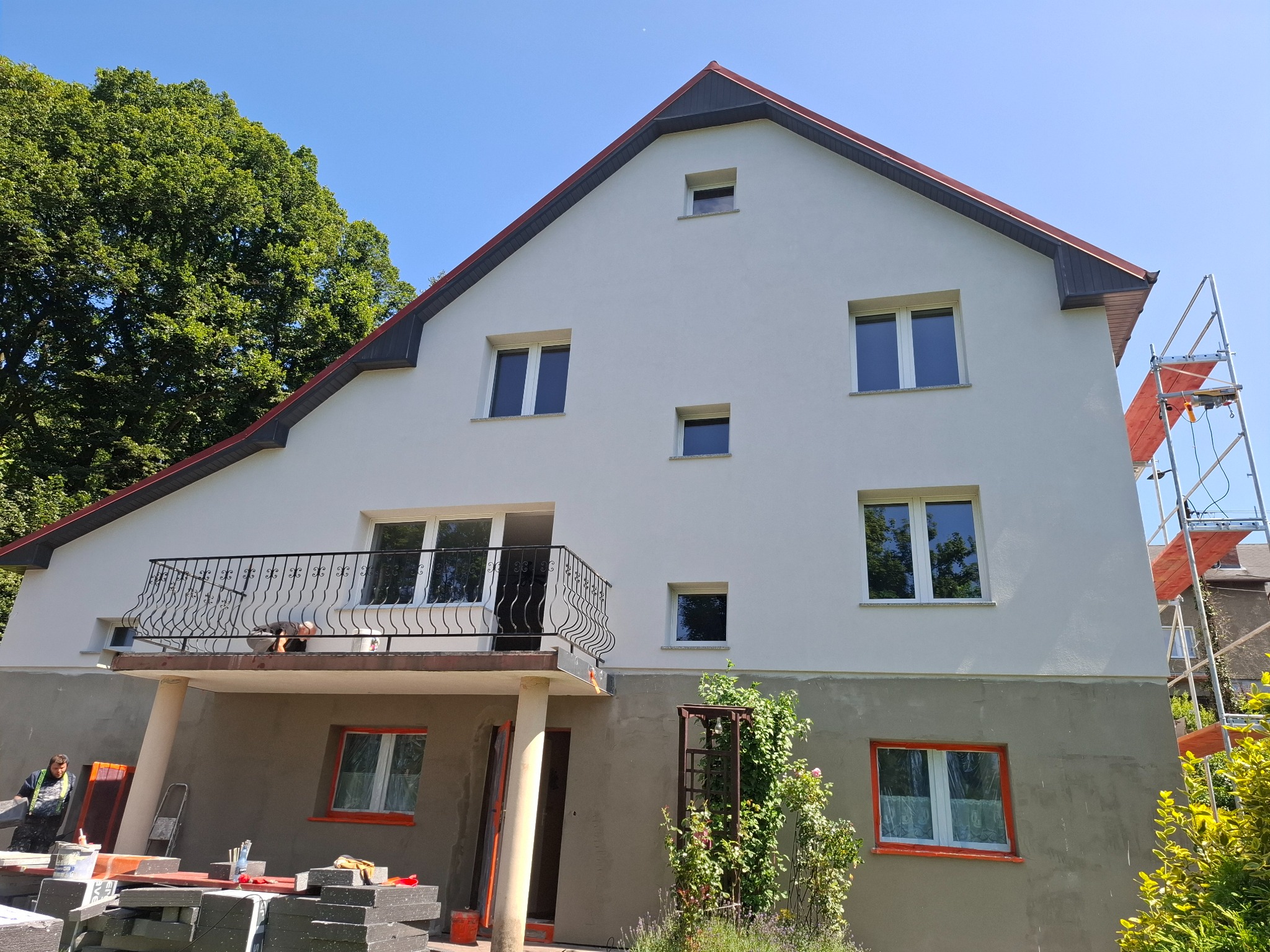 Dwupiętrowy dom jednorodzinny z balkonem w trakcie prac elewacyjnych; dolna część budynku otynkowana na szaro, górna pomalowana na biało, widoczne rusztowanie i materiały budowlane.
