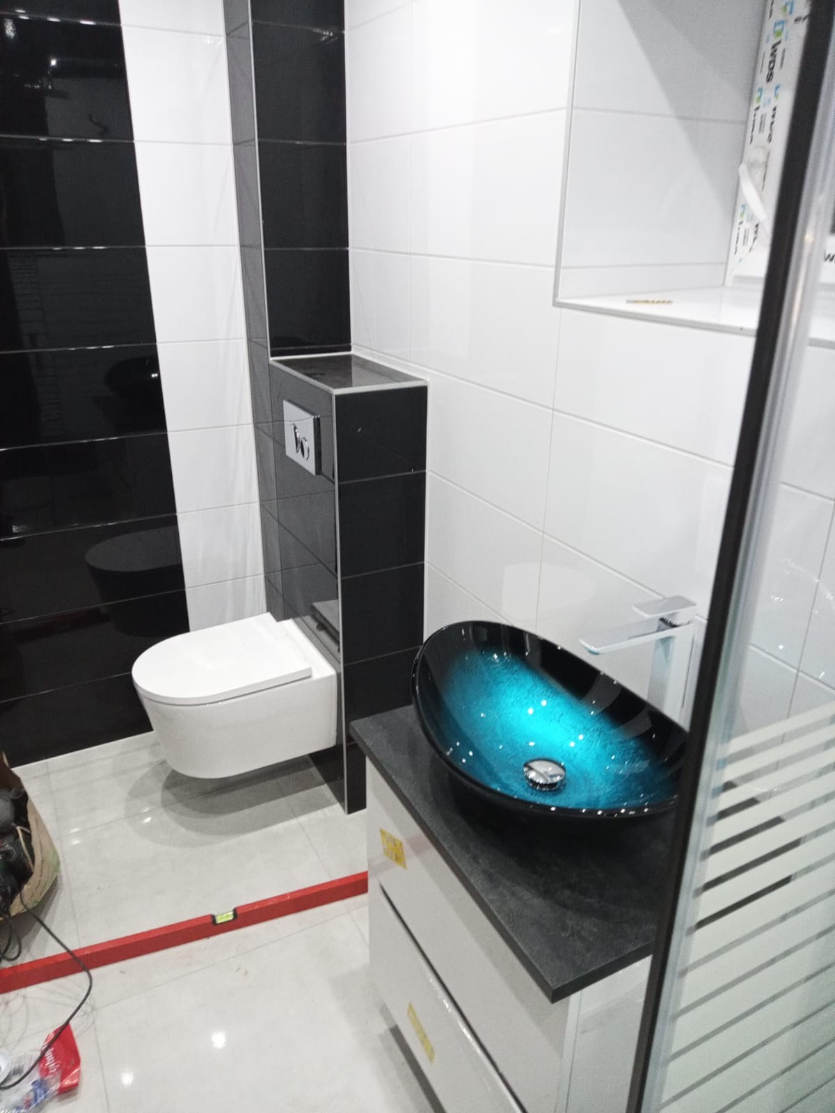 Nowoczesna łazienka z czarno-białymi płytkami, podwieszaną toaletą i umywalką w kolorze turkusowym na szafce z szufladami, widoczny poziomica na podłodze.