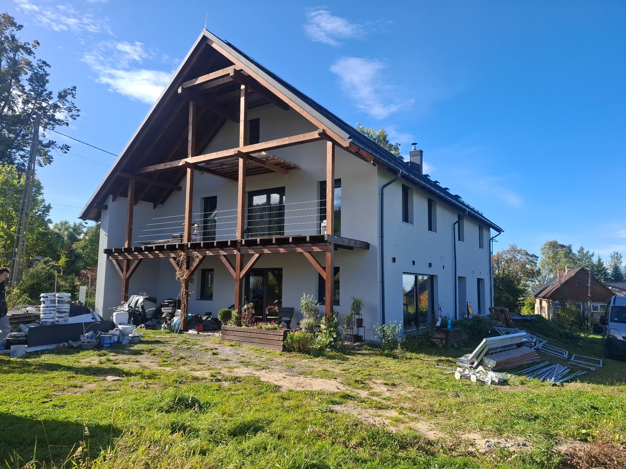 Dwukondygnacyjny dom z jasną elewacją, drewnianą konstrukcją dachu i balkonami, widoczny w słoneczny dzień z elementami placu budowy wokół.