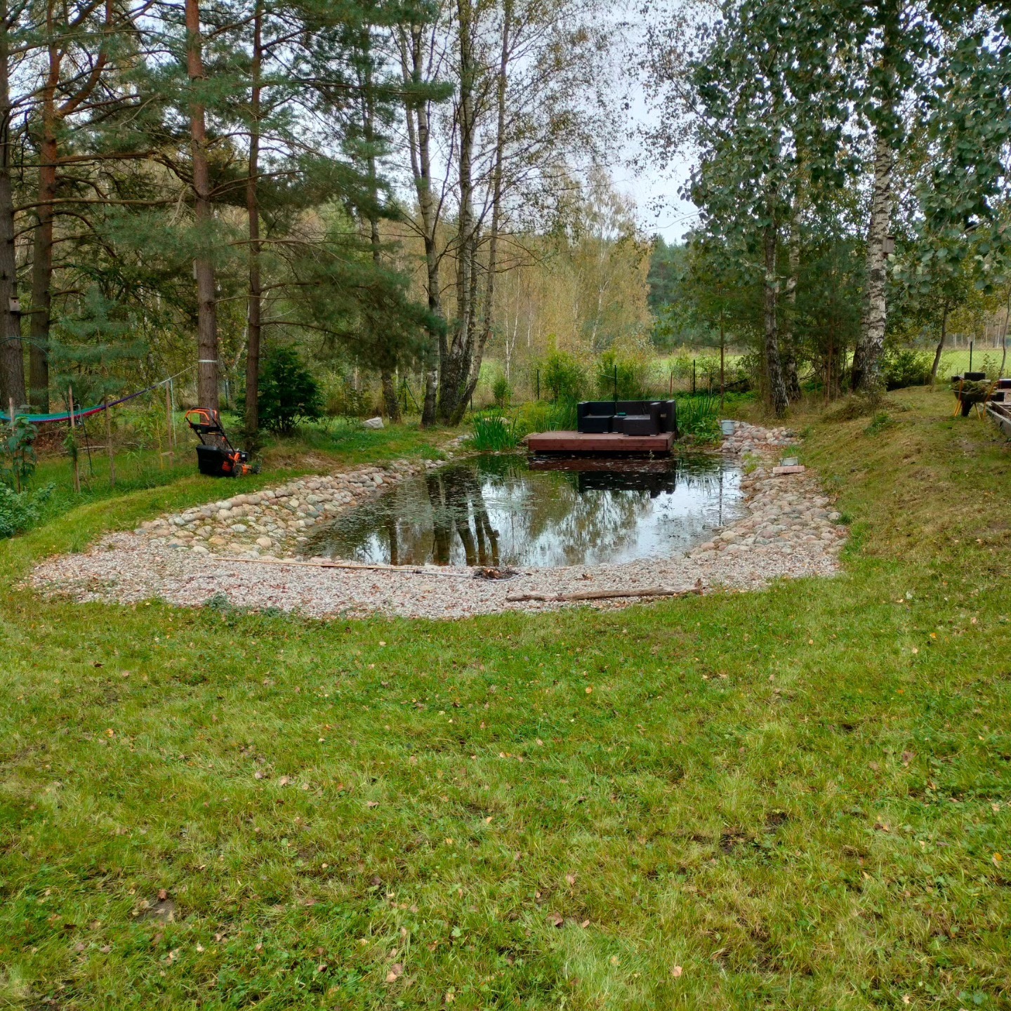 Oczko wodne z drewnianym pomostem i meblami ogrodowymi w otoczeniu drzew i trawnika w Zbychowie, widoczna kosiarka do trawy.
